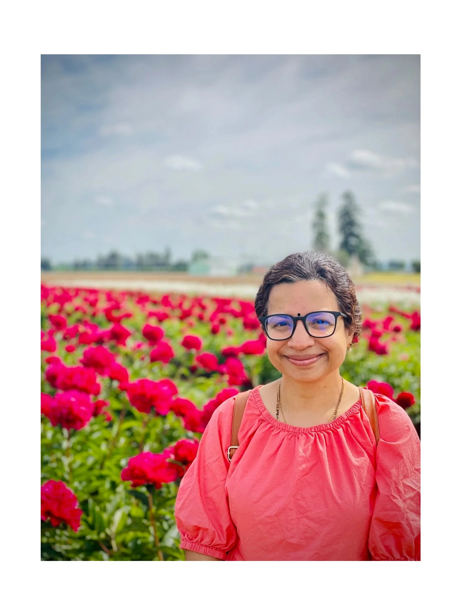 A woman smiling in a red top standing in a vibrant flower field with pink and red flowers, under a partly cloudy sky.