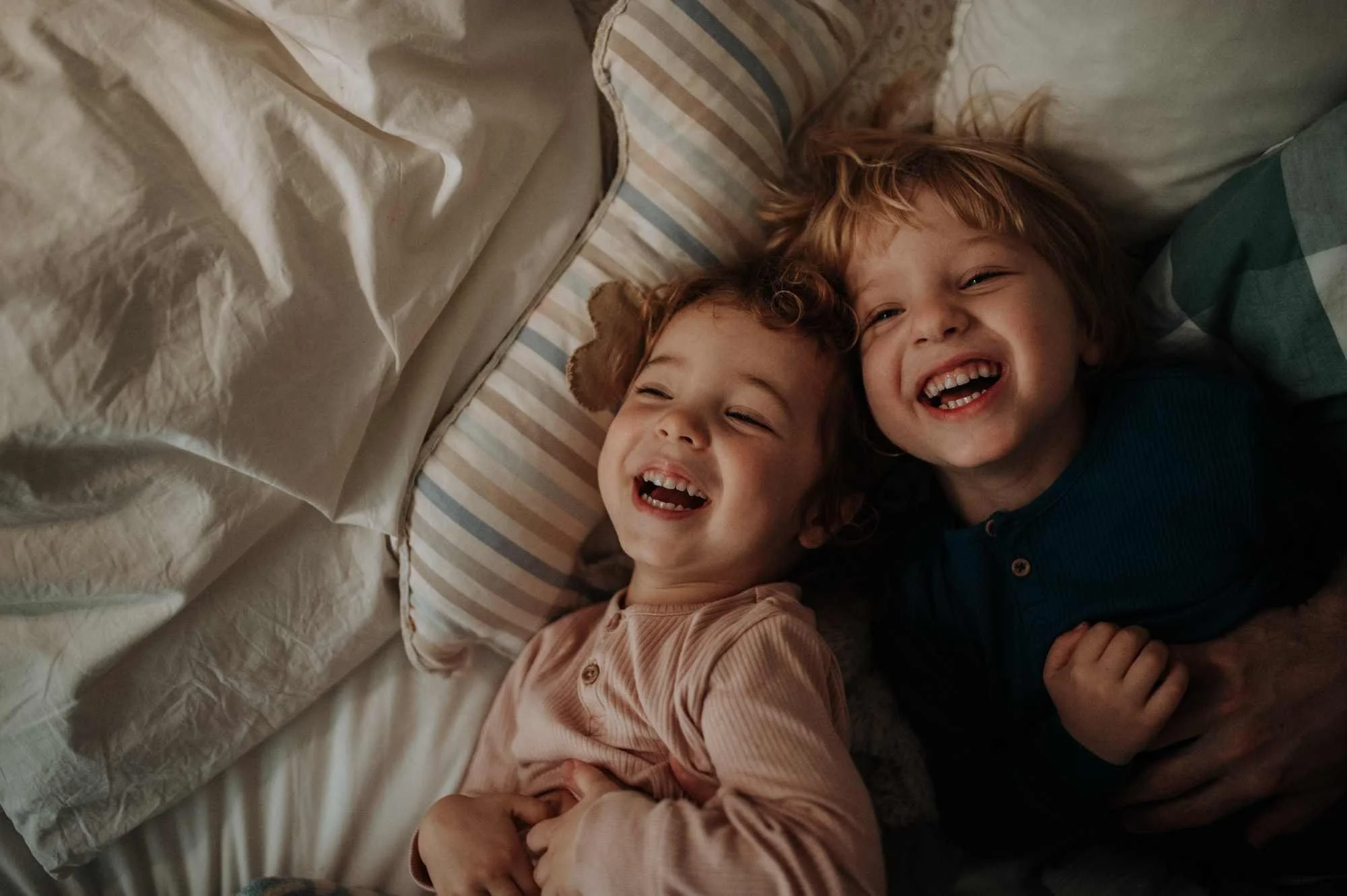 Two young children lying in bed, smiling and laughing, surrounded by pillows and blankets.
