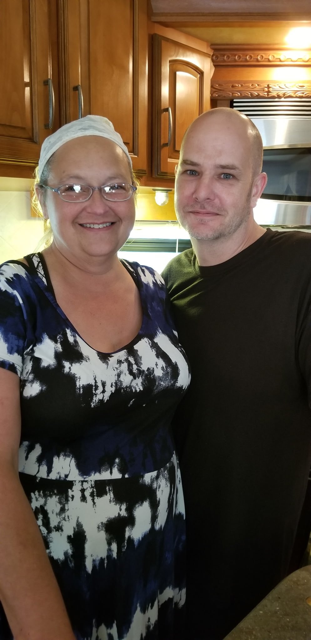 A woman and a man standing in a kitchen taking a selfie. The woman is smiling, wearing glasses, a white head covering, and a black and white patterned dress. The man is standing close, smiling, and wearing a black t-shirt. The kitchen background has wooden cabinets and a stainless steel microwave.