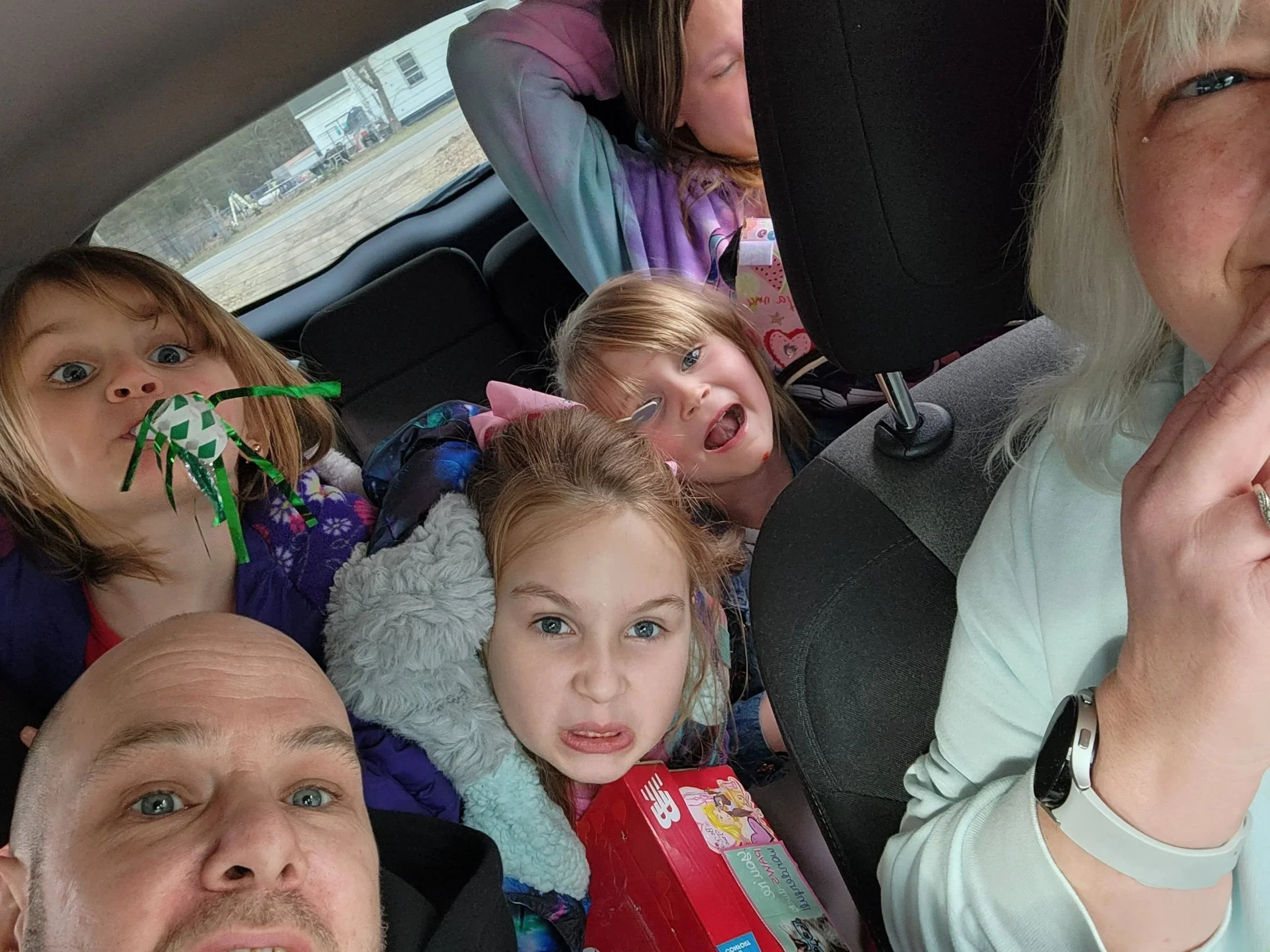 A group of five children and one adult inside a car. Four children are in the back seat, with one holding a toy pipe cleaner. The adult is in the front passenger seat, partially visible. The children are making silly faces.