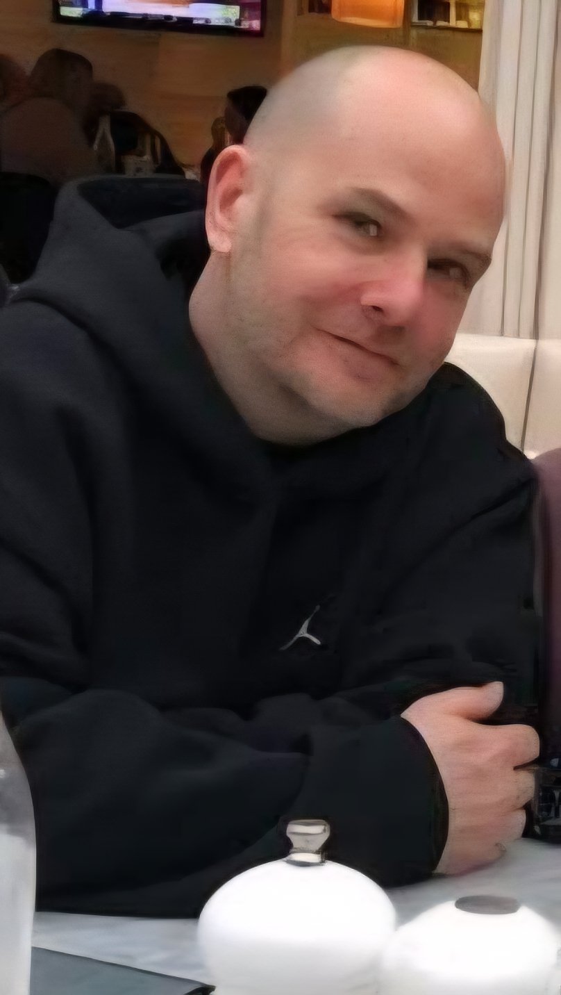 A bald man with a light complexion, smiling slightly, sitting at a table in a restaurant or cafe. He is wearing a black hoodie and resting his right arm on the table. In the background, other diners and a TV screen are visible.