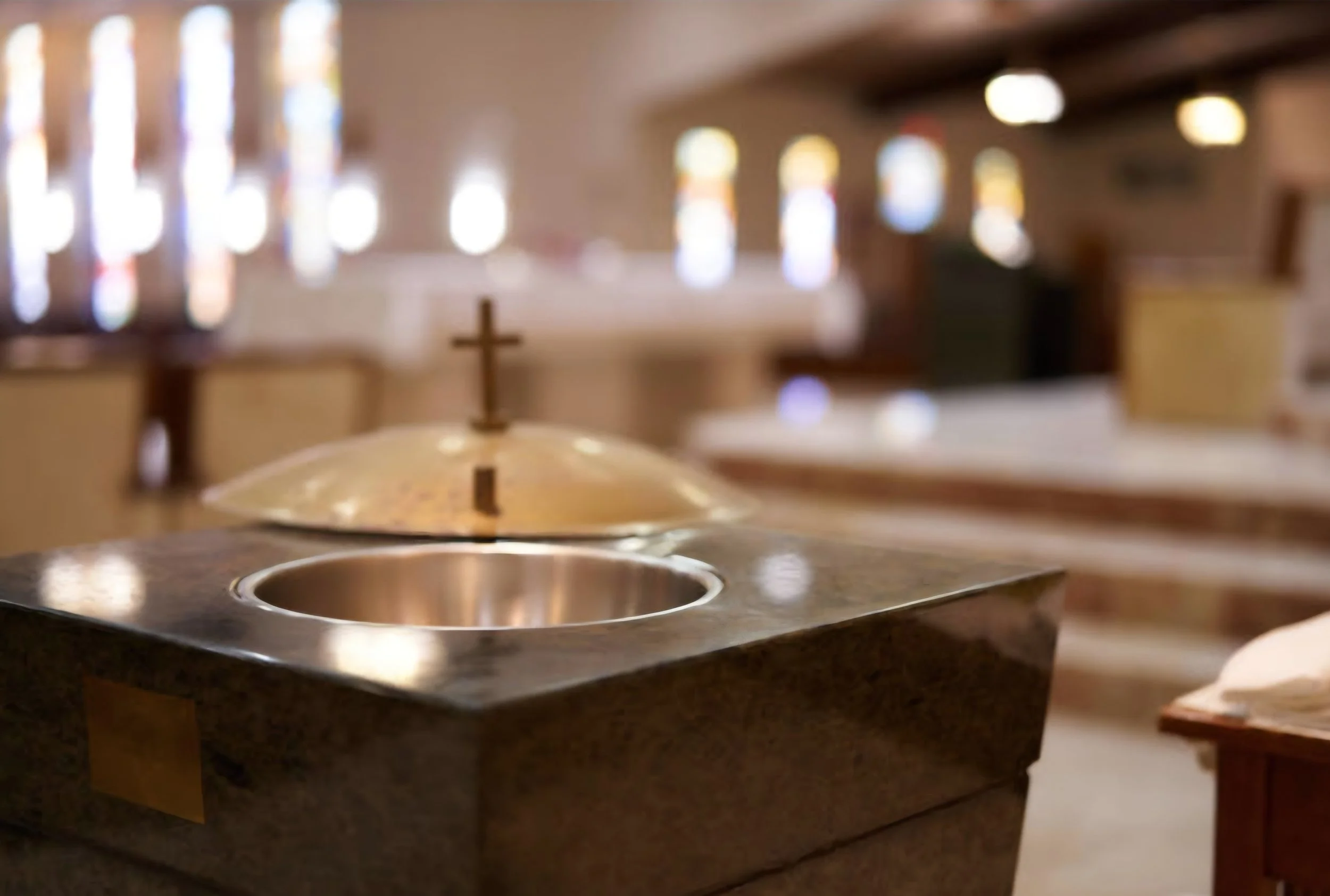 Liturgical chalice and ciborium with a cross on top inside a church or chapel with stained glass windows and wooden furnishings in the background.