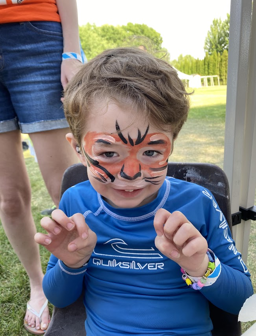 Young boy with face paint resembling a tiger, wearing a blue Quiksilver shirt, sitting on a chair outdoors, smiling and making paws with his hands.