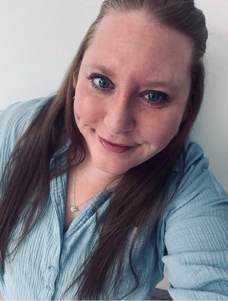 A woman with long brown hair, blue eyes, and a nose piercing, smiling and wearing a light blue shirt, taking a selfie against a plain white wall.