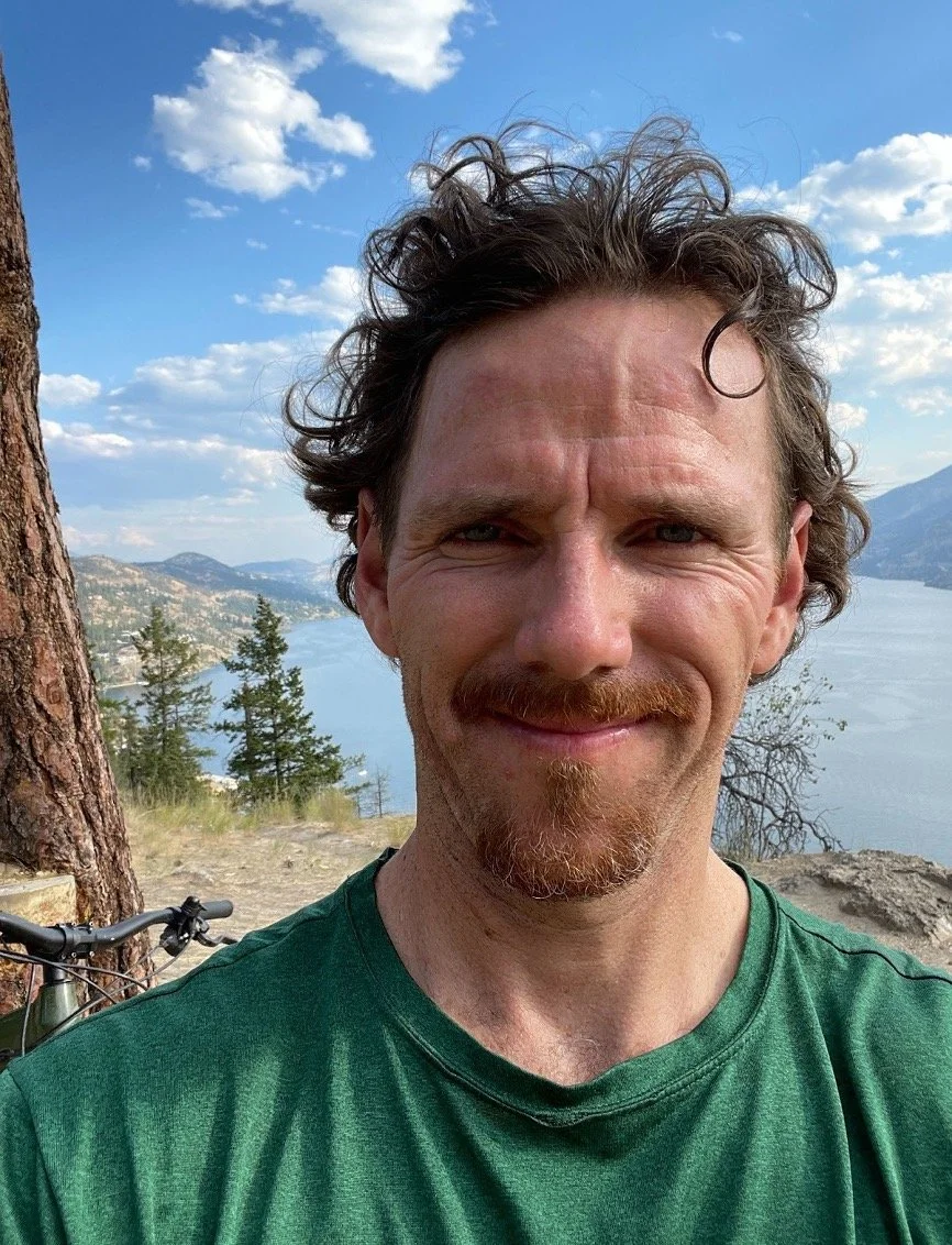 A man with curly hair and a beard, smiling, standing outdoors near a body of water with mountains and trees in the background on a partly cloudy day.