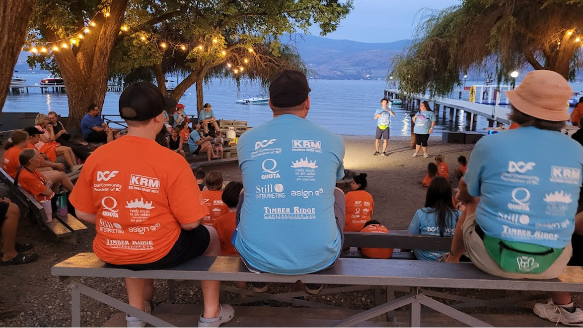 People sitting on benches and standing near a lakeside with trees and string lights, watching a performance on the beach, with mountains and a pier in the background.