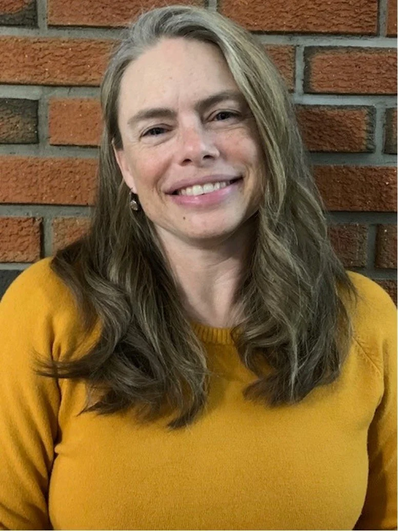 A woman with shoulder-length wavy brown hair, smiling, wearing a mustard yellow sweater, standing in front of a brick wall.