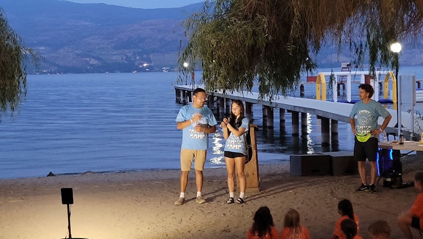 Group of three people speaking on a beach at camp, with children sitting in front. There is a dock and water in the background, trees overhead, and a table with equipment on the right.