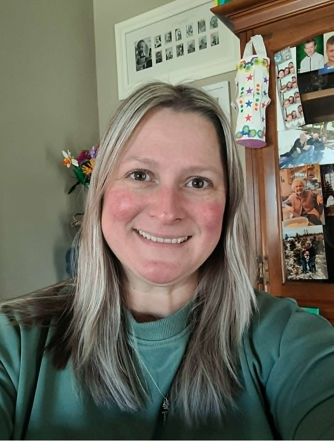 A woman with blonde hair and freckles smiling indoors, with a wallpaper of family photos and a colorful gift bag in the background.