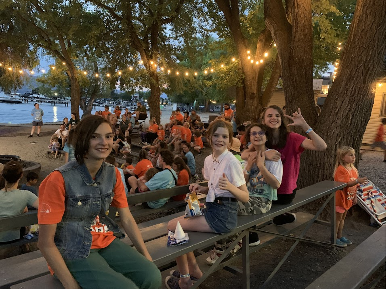 Group of children and teenagers enjoying an outdoor gathering by a waterfront at dusk, sitting on benches under string lights and large trees.