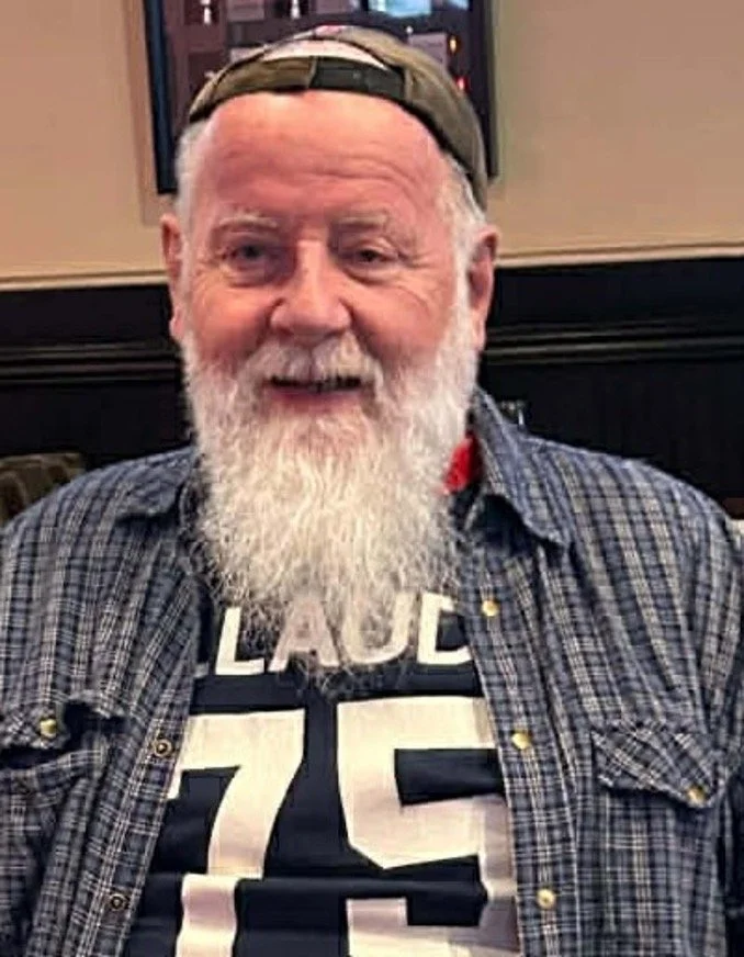 An elderly man with a white beard and hair, wearing a baseball cap backwards, plaid shirt, and a sports jersey with the name 'LADUE' and the number '75'.