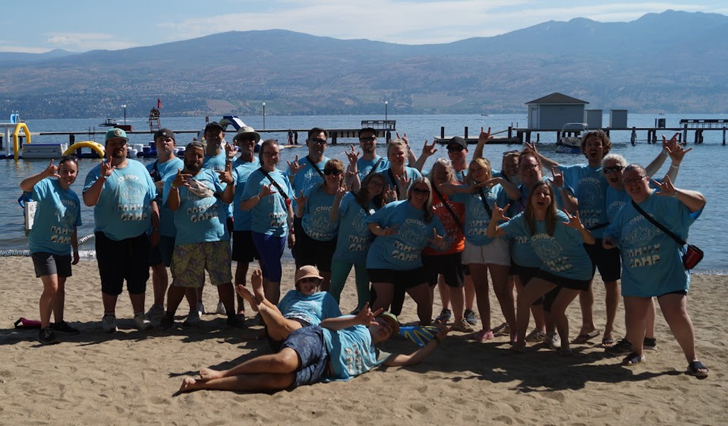 Group of people on a sandy beach by a lake, posing for a photo with mountains in the background, several making peace signs, some wearing blue matching shirts.