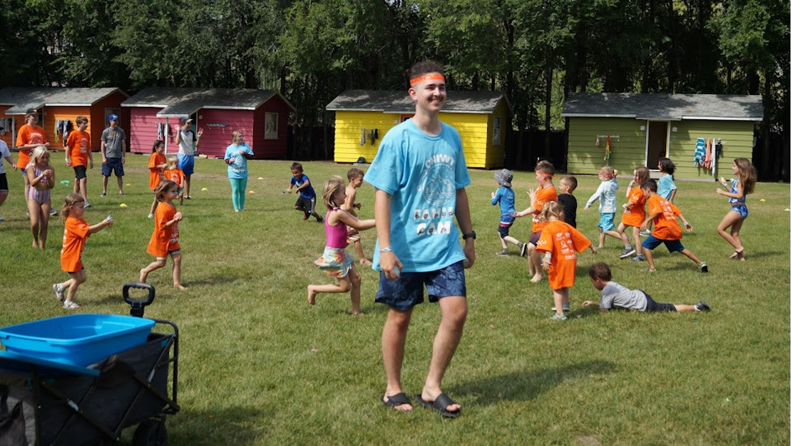 A group of children and teenagers playing outdoor games on a grassy field at summer camp, with colorful small cabins in the background.