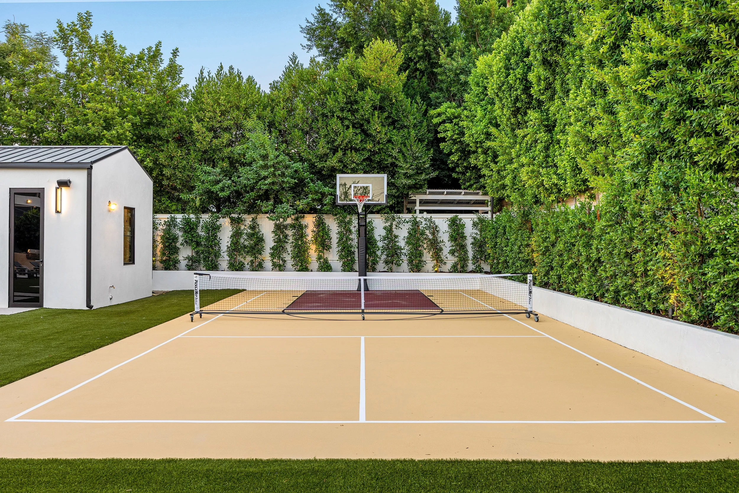 Empty backyard with a tennis court and basketball hoop, surrounded by tall green trees and a white fence, with a small modern building on the left side.