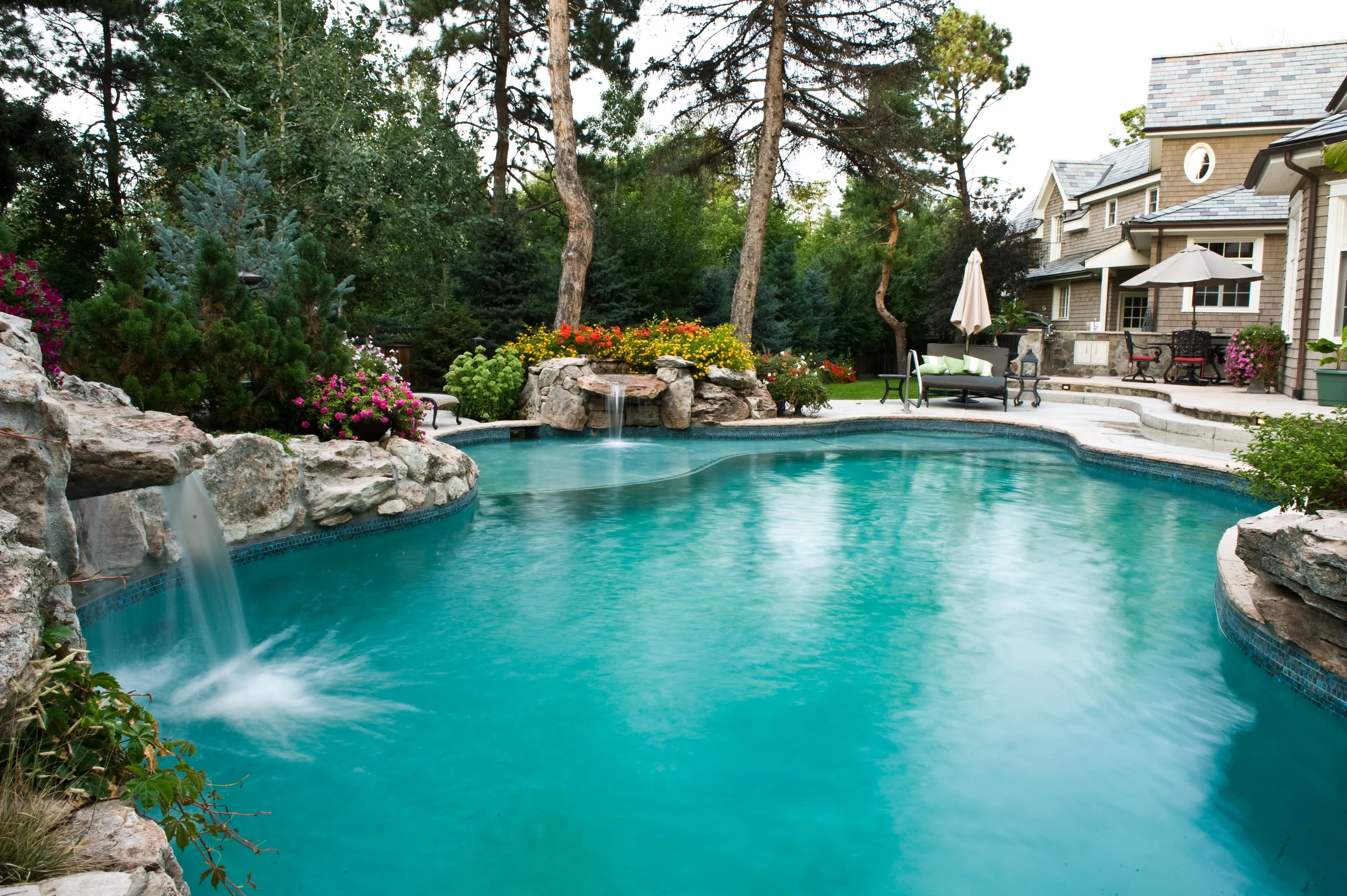 Backyard with a swimming pool, surrounded by rocks, flowers, and trees, with outdoor furniture and a house in the background.