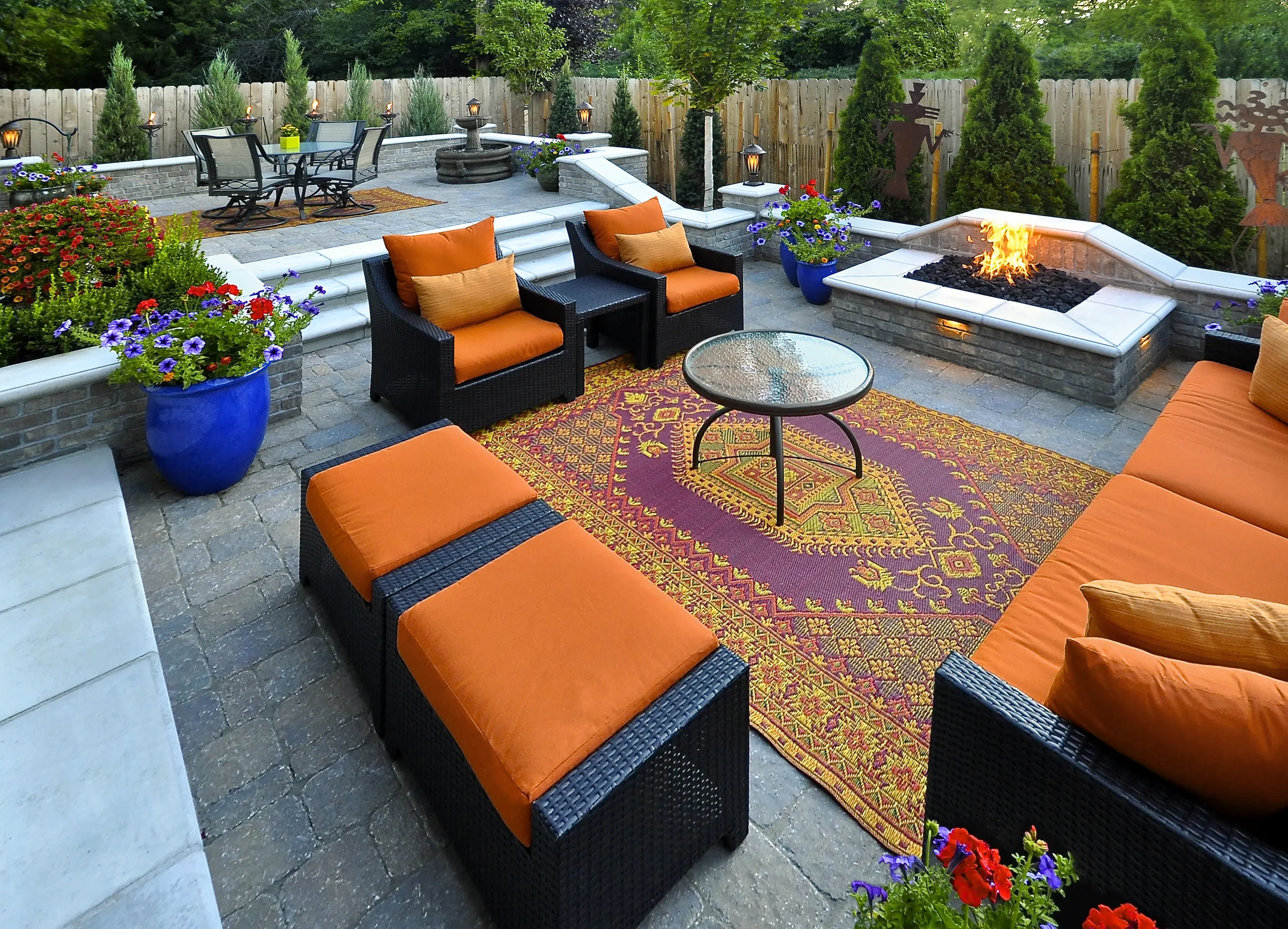 An outdoor patio area features black wicker furniture with bright orange cushions, a colorful patterned rug, potted flowers, and a modern gas fire pit on a stone deck, surrounded by a wooden fence and lush greenery.