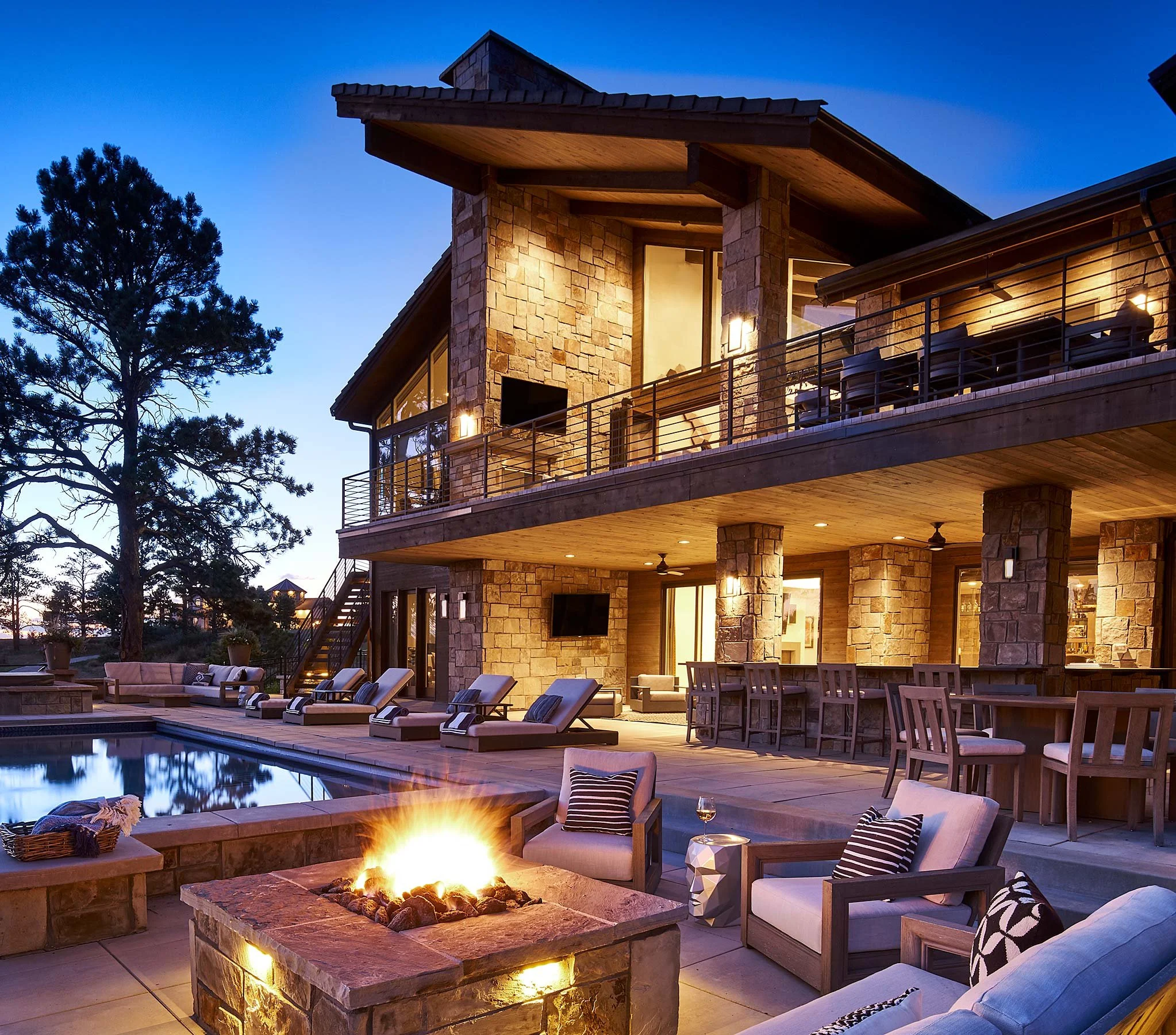 Luxury house with stone exterior featuring multiple balconies illuminated at dusk, with a backyard patio pool area including sun loungers, a fire pit with a fire, seating with pillows, a bar area, and a view of tall trees.