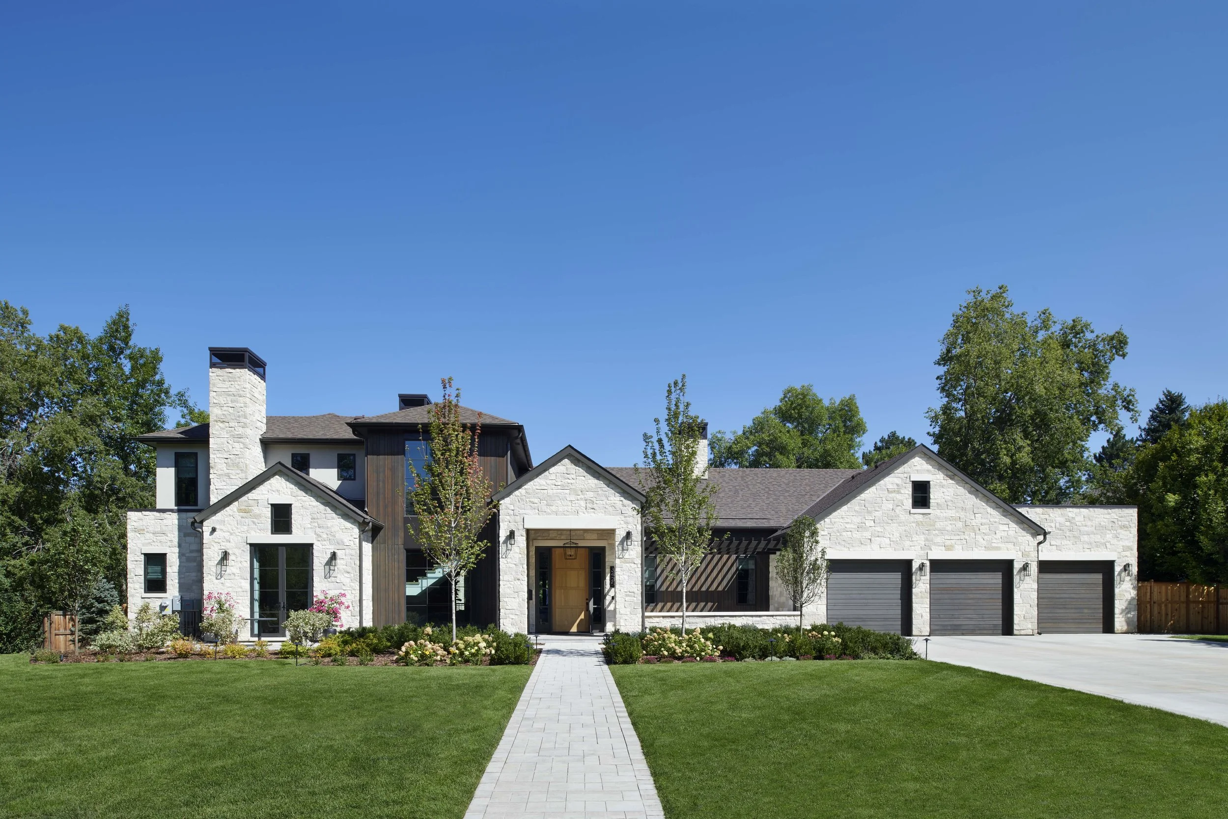 Modern two-story house with white stone exterior, large windows, and three black garage doors. Well-maintained front yard with green lawn, trees, and flower beds, under a clear blue sky.