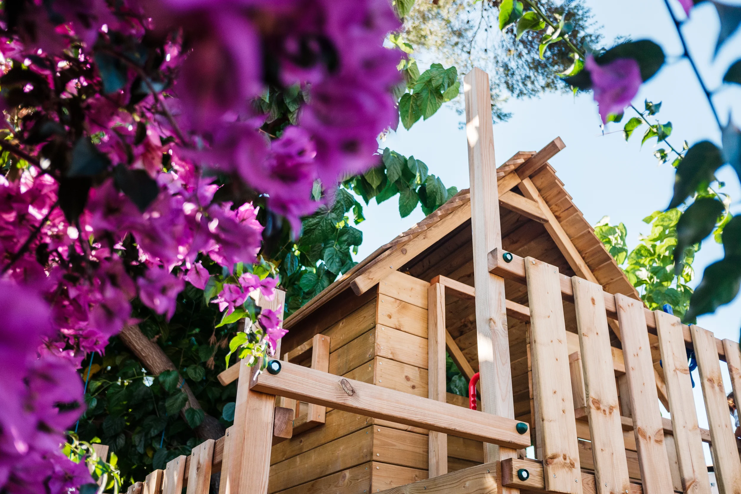 A wooden treehouse surrounded by purple flowers and green foliage with a clear blue sky in the background.