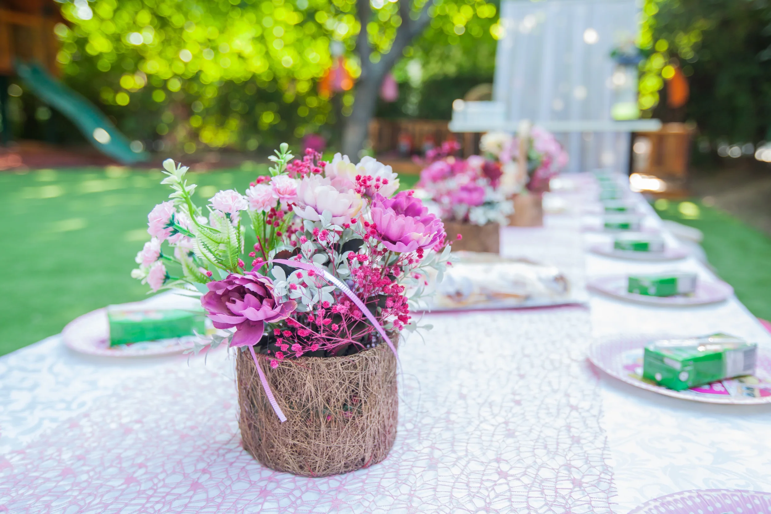 Close-up of a floral centerpiece with pink and purple flowers on a decorated table set for an outdoor gathering or party, with additional floral arrangements and chairs in the background under trees.
