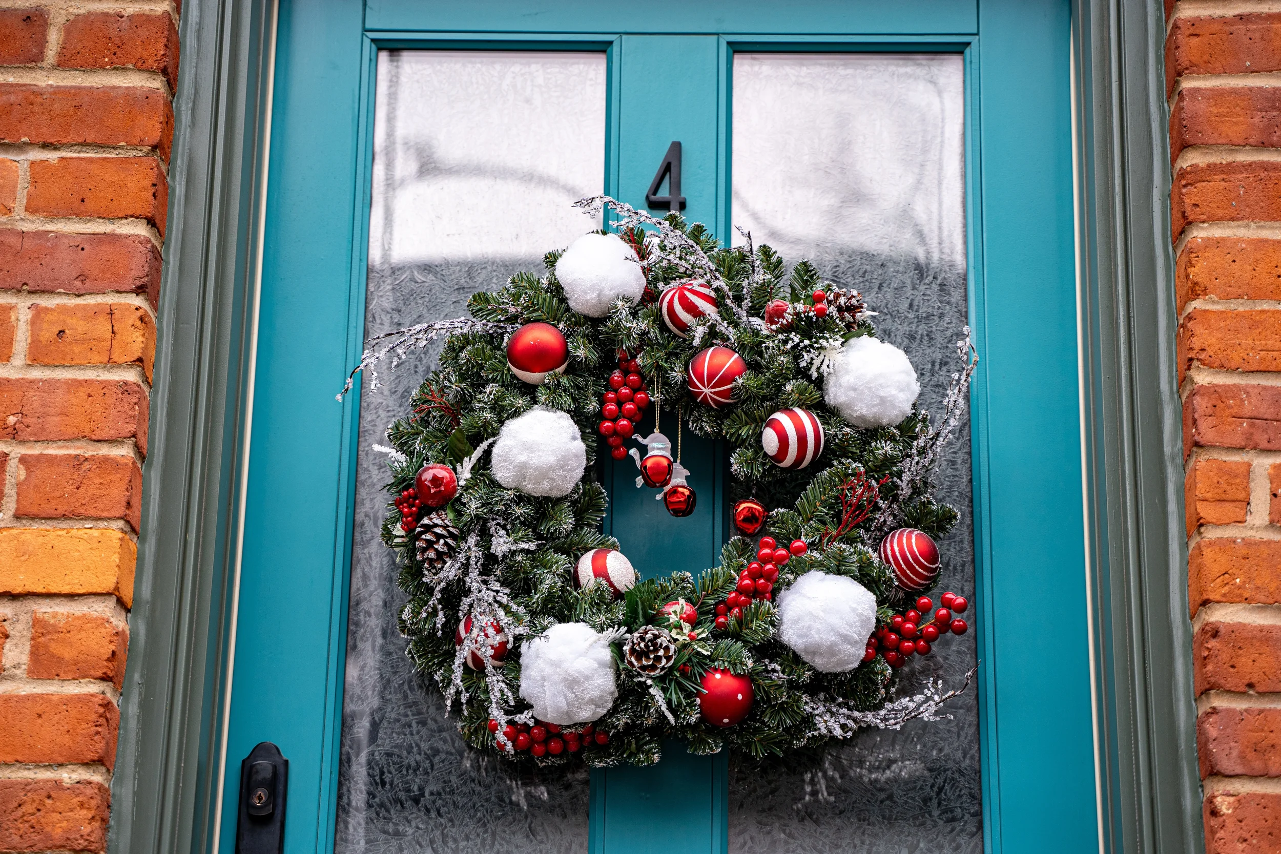 Christmas wreath with red and white ornaments, pinecones, and snow decorations hanging on a teal front door with the number 4.