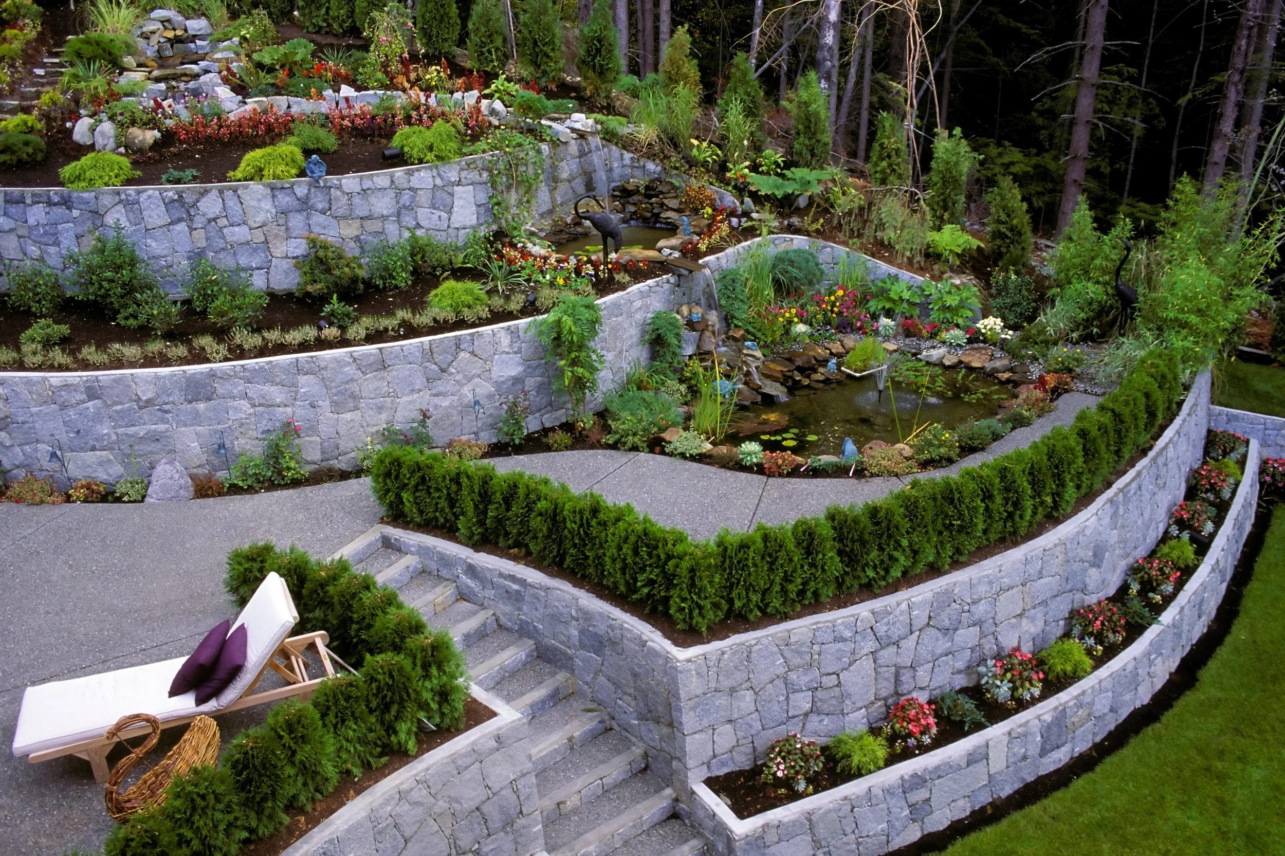 Multi-tiered garden with stone retaining walls, lush green plants, colorful flowers, and water features including a pond and small waterfalls. A lounge chair with cushions is on a paved patio area.