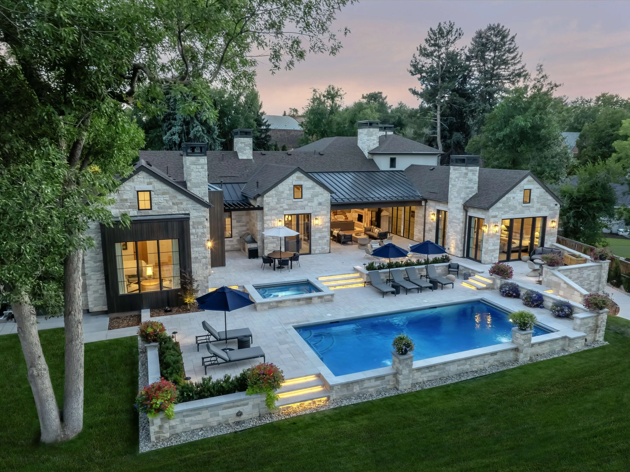 A luxurious modern backyard with a swimming pool, hot tub, outdoor seating area, umbrellas, and a large stone house with multiple chimneys at dusk.