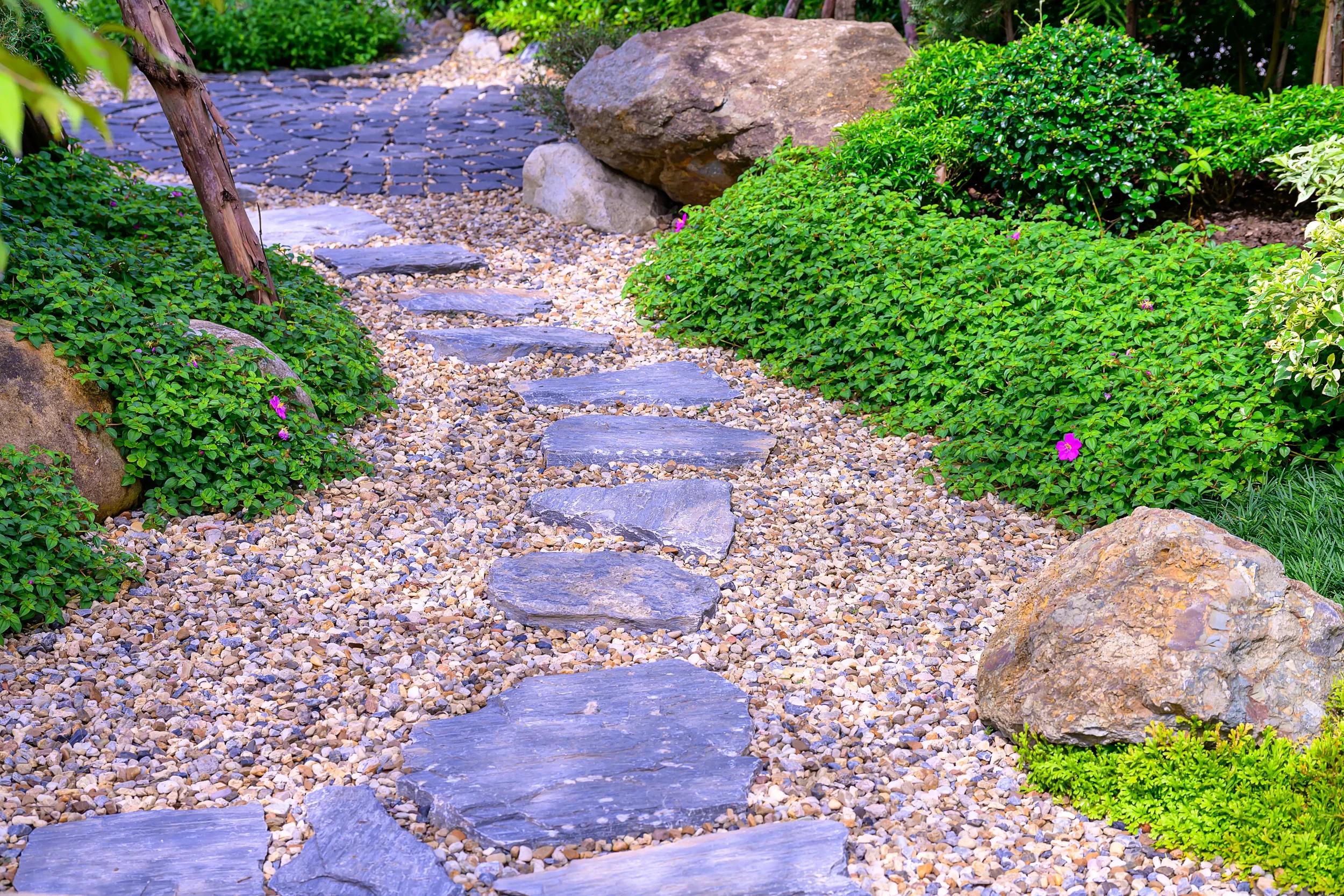 A garden pathway made of irregular flat stones set in small pebbles, surrounded by lush green bushes, small plants, and large rocks.