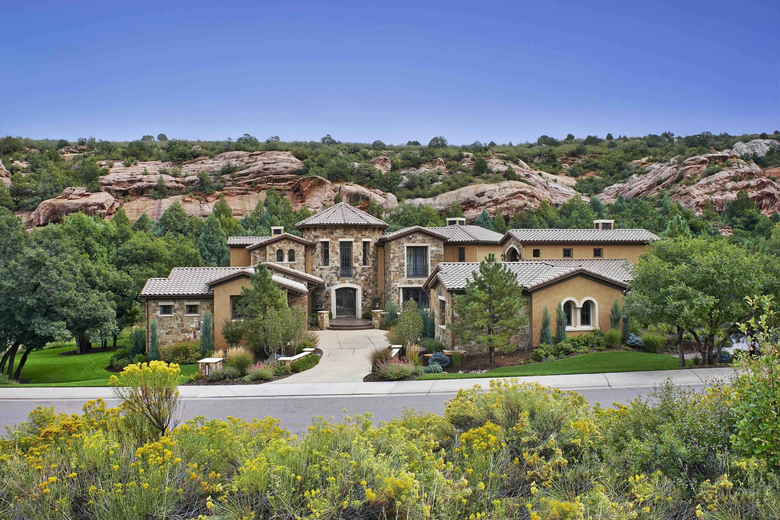 Large luxurious house with stone and stucco exterior, surrounded by lush landscaping, at the foothills of rocky mountains under a blue sky.