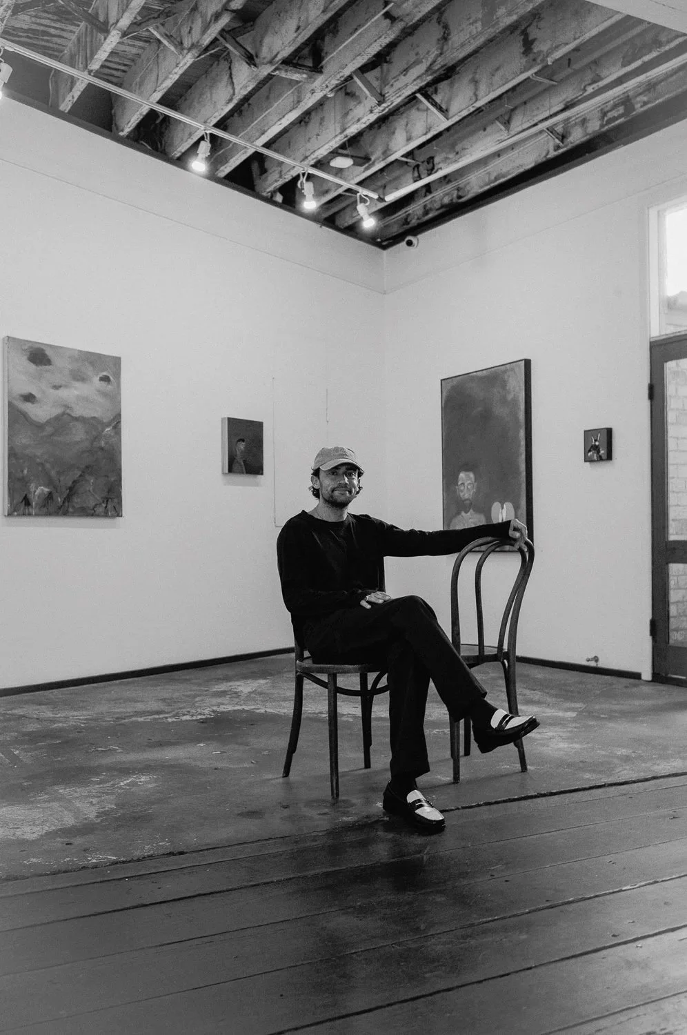 A man sitting on a wooden chair in an art gallery with painting artworks on the wall behind him, black and white photograph.