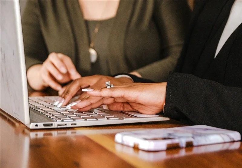 Two women are sitting at a table with a laptop and a smartphone, one woman is typing on the laptop keyboard.
