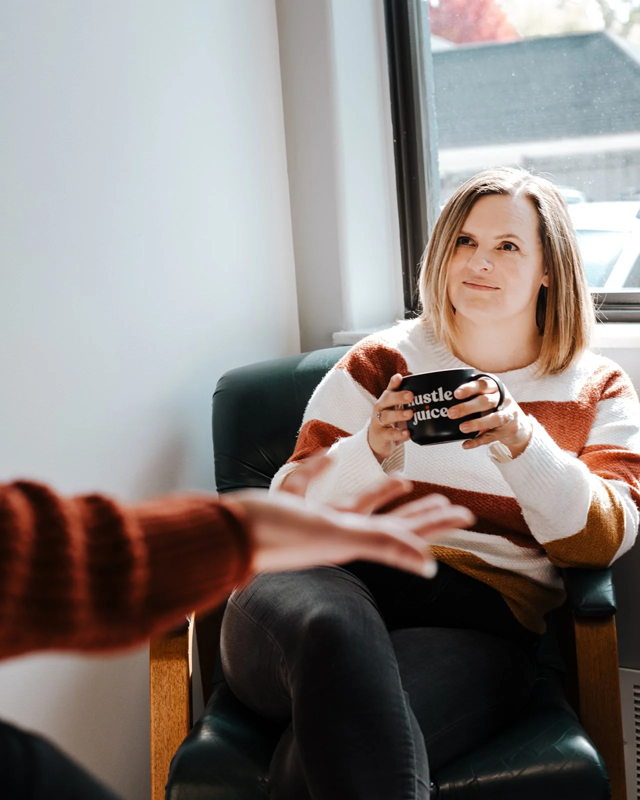 Danielle Allison sitting in a green armchair holding a black mug, actively listening while engaged in conversation, next to a window.