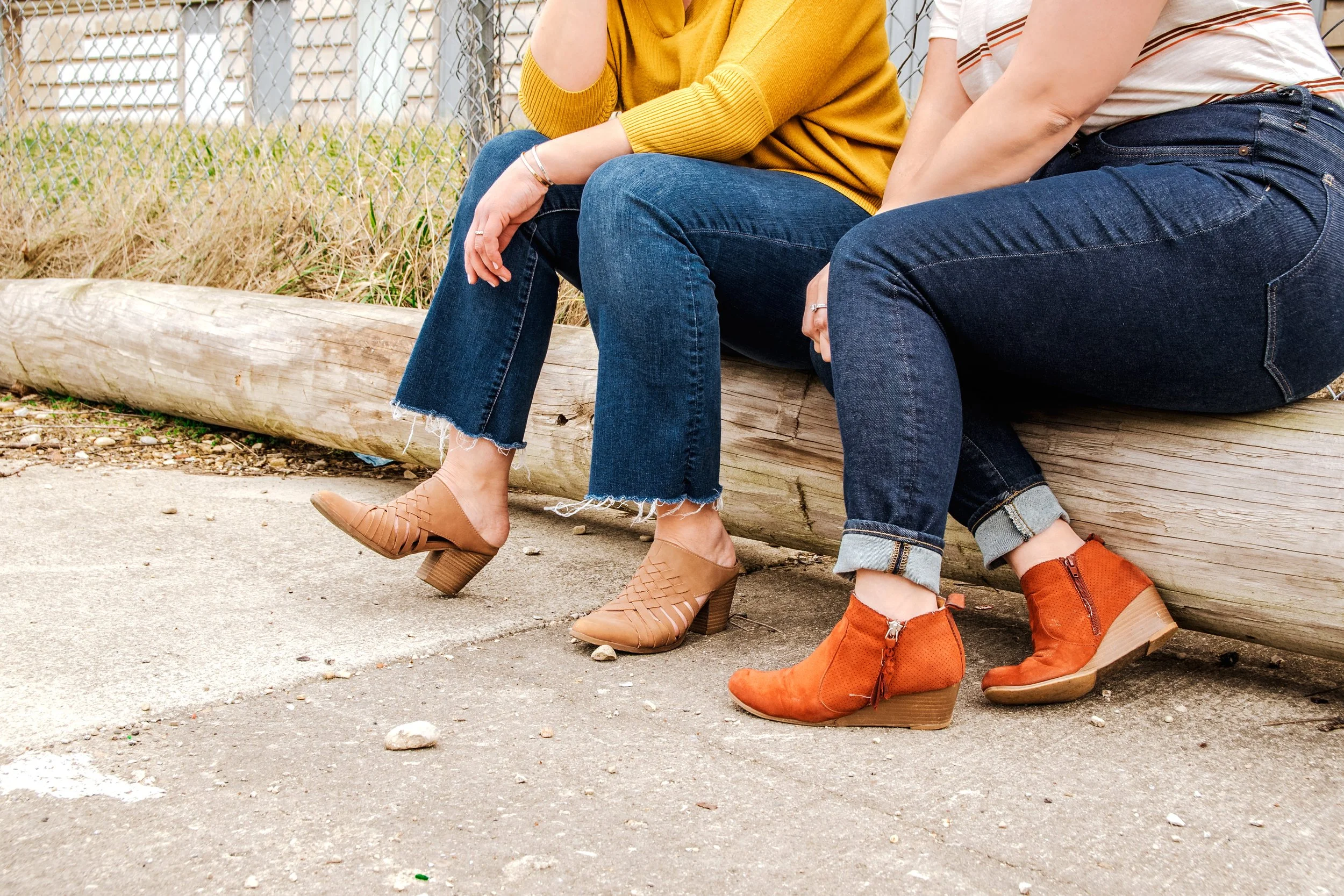 Two women sitting on a wooden log, showing only their legs and feet, on a concrete path with grass and a chain-link fence in the background. They are wearing jeans and stylish ankle boots.