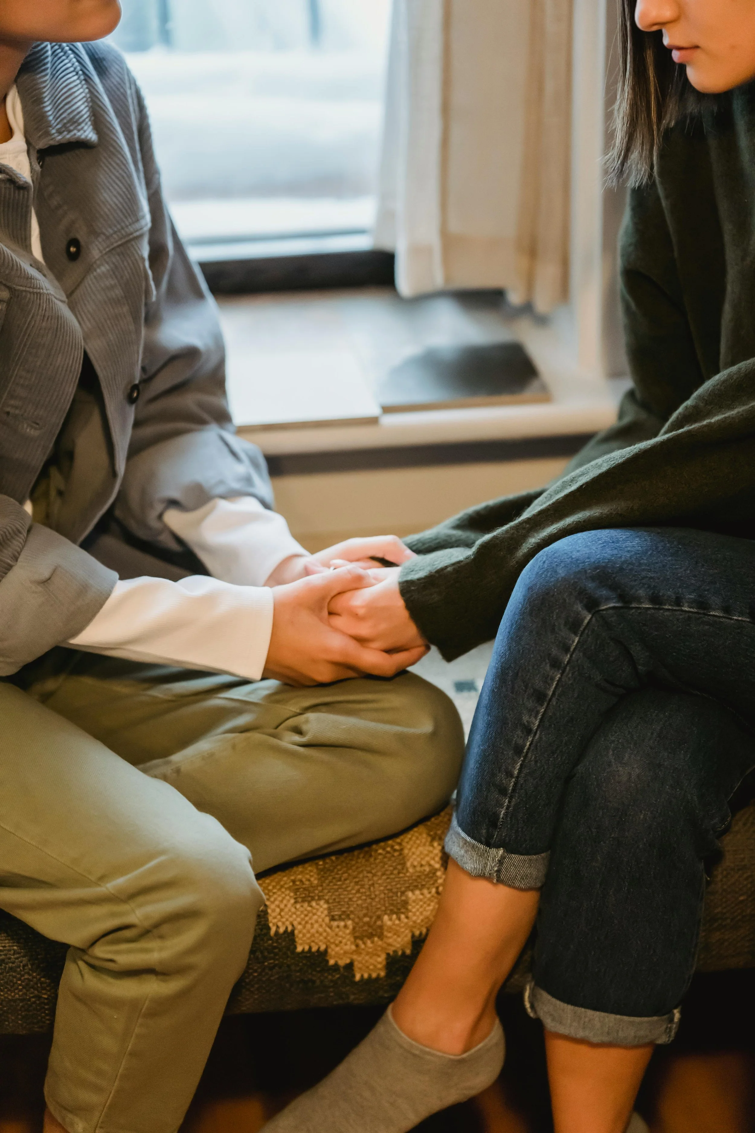 Two people sitting on a bench by a window holding hands in a show of support.