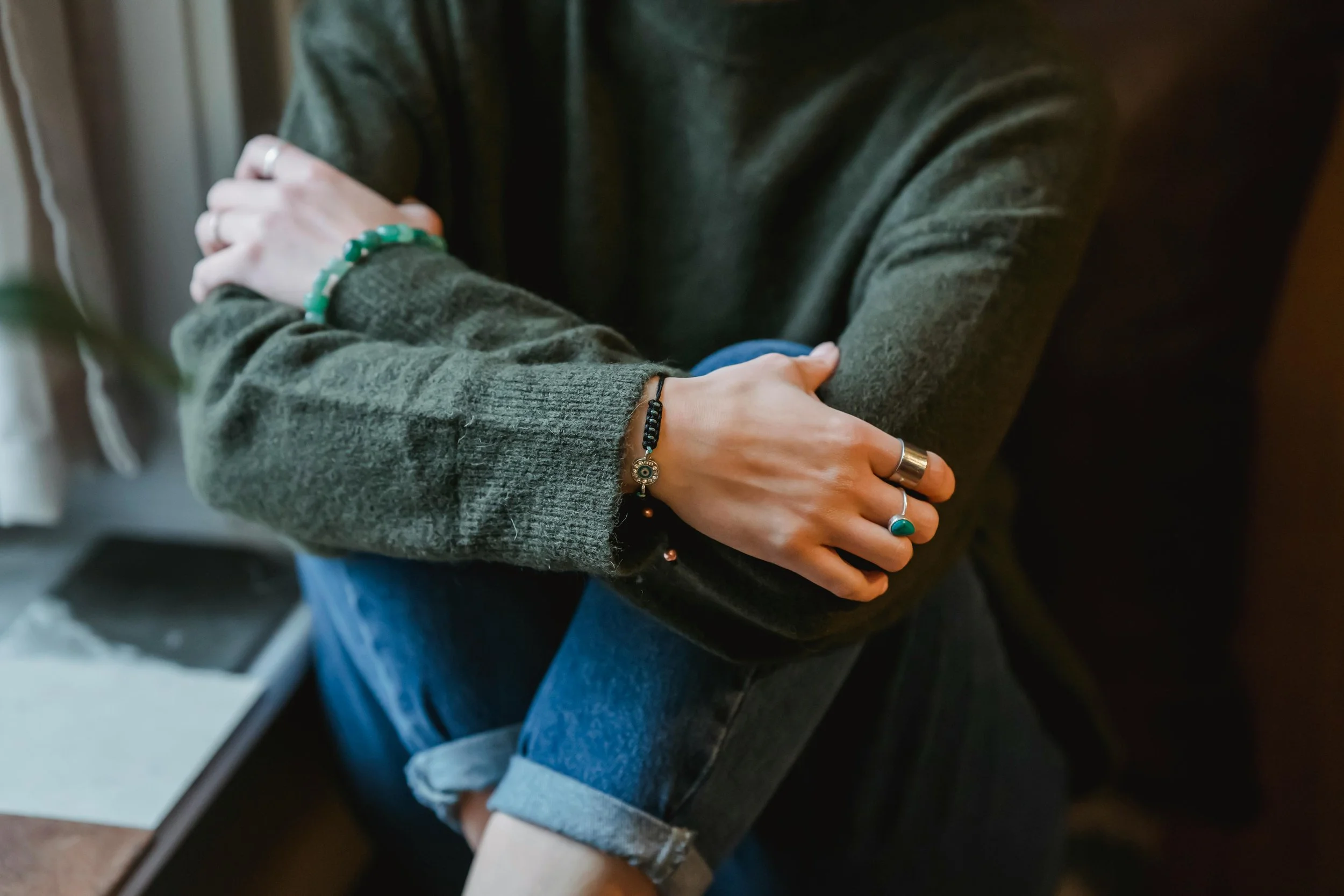 Person sitting with arms crossed, wearing a green sweater, blue jeans, and multiple rings and bracelets.