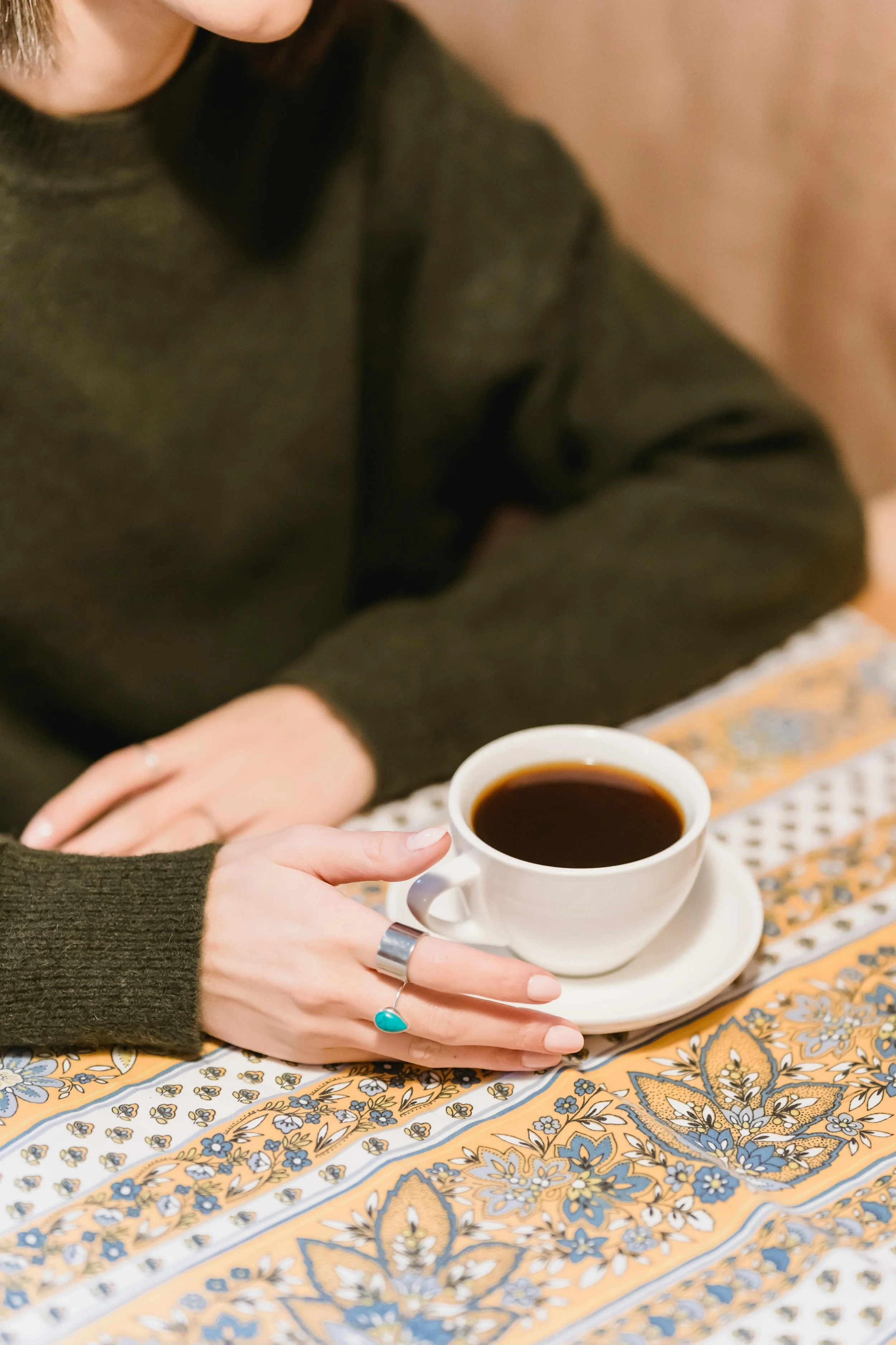 A person with light skin, holding a white coffee cup and saucer, sitting at a table with a colorful patterned tablecloth. The person is wearing a dark green sweater and a silver ring with a turquoise stone on their hand. The person's face is not visible in the image.