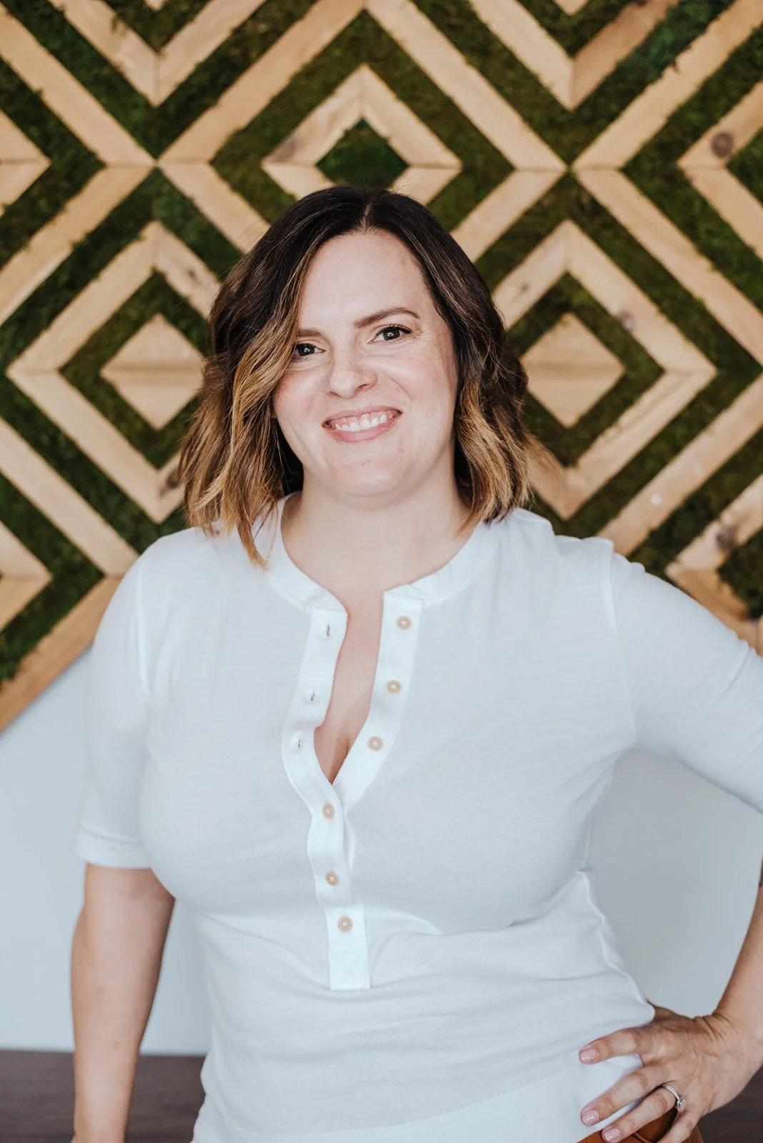 Danielle Allison with shoulder-length wavy hair, wearing a white shirt with a buttoned neckline, smiling and standing in front of a geometric wooden wall with green moss accents.