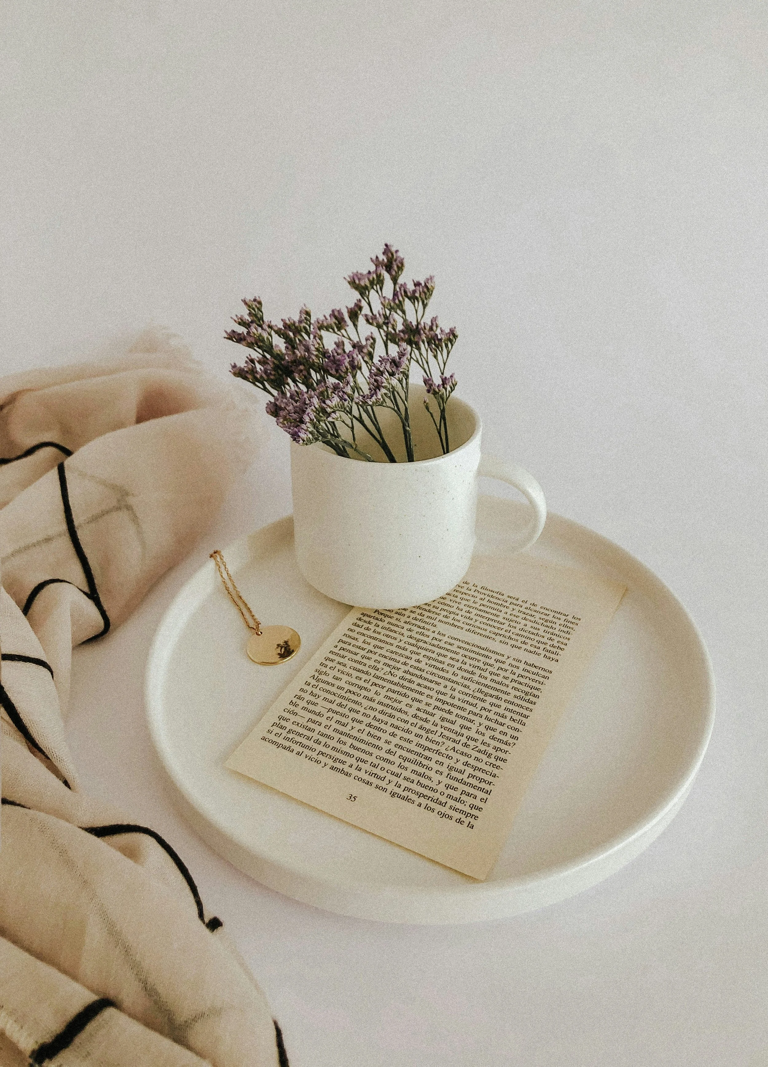 A white tray holds an open book, a white mug with purple flowers, and a gold necklace with a round pendant. A beige cloth with black lines is partially visible on the left.