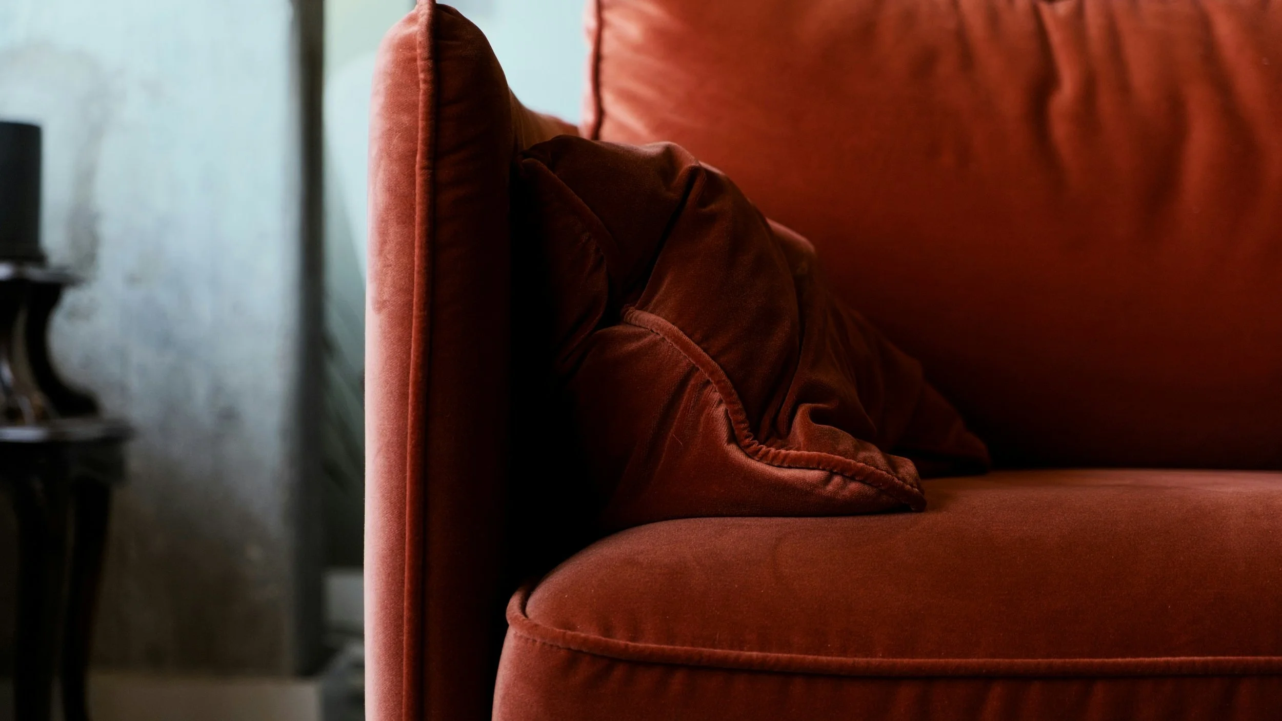 Close-up of a velvet armrest on a burnt orange sofa with matching cushions. A dark metal side table is partially visible on the left.