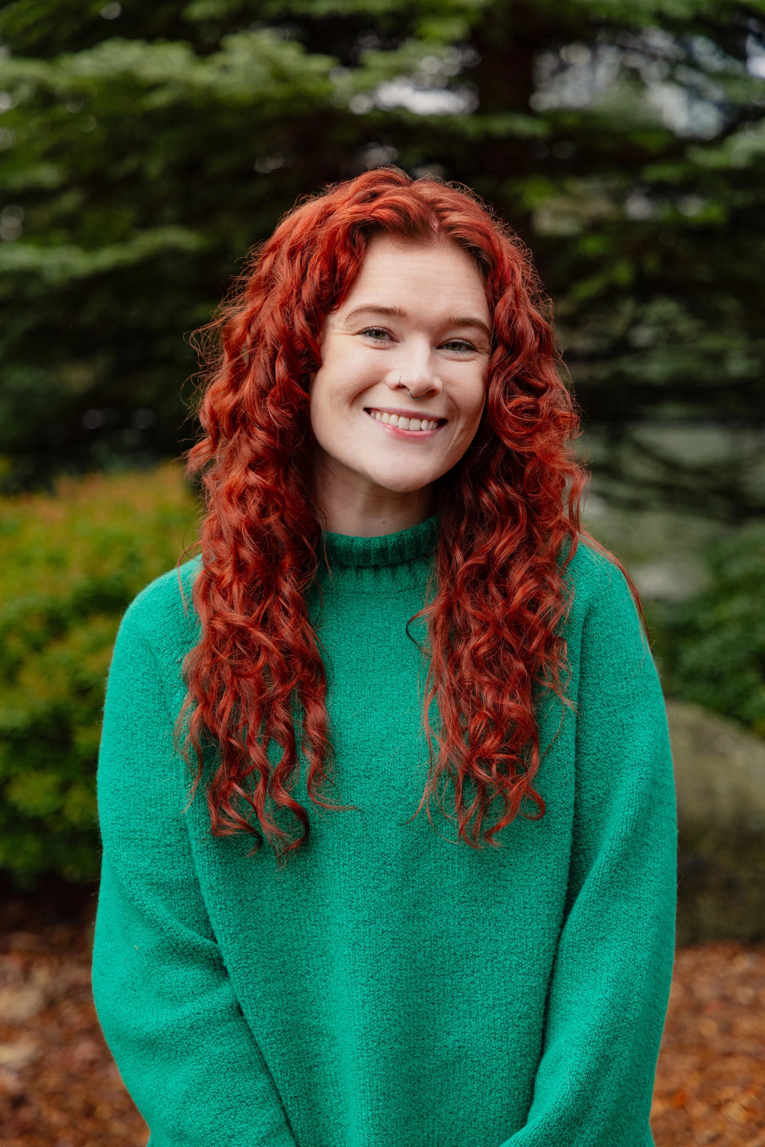 A young woman with long, curly red hair and a septum piercing, smiling outdoors in front of green trees and foliage, wearing a teal sweater.