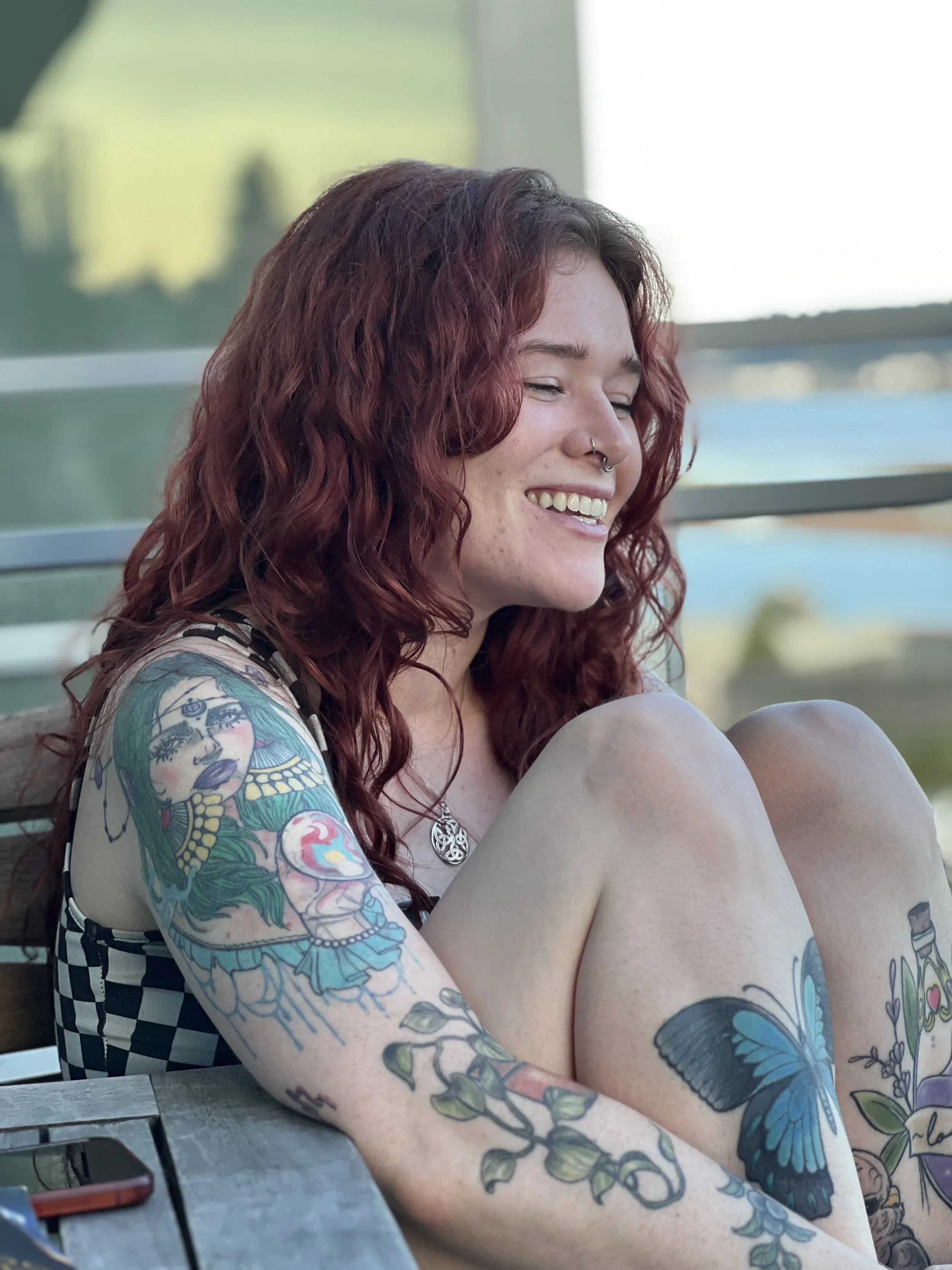 A young woman with curly red hair and tattoos on her arms and legs, sitting outdoors with a big smile, near a window and a scenic background.