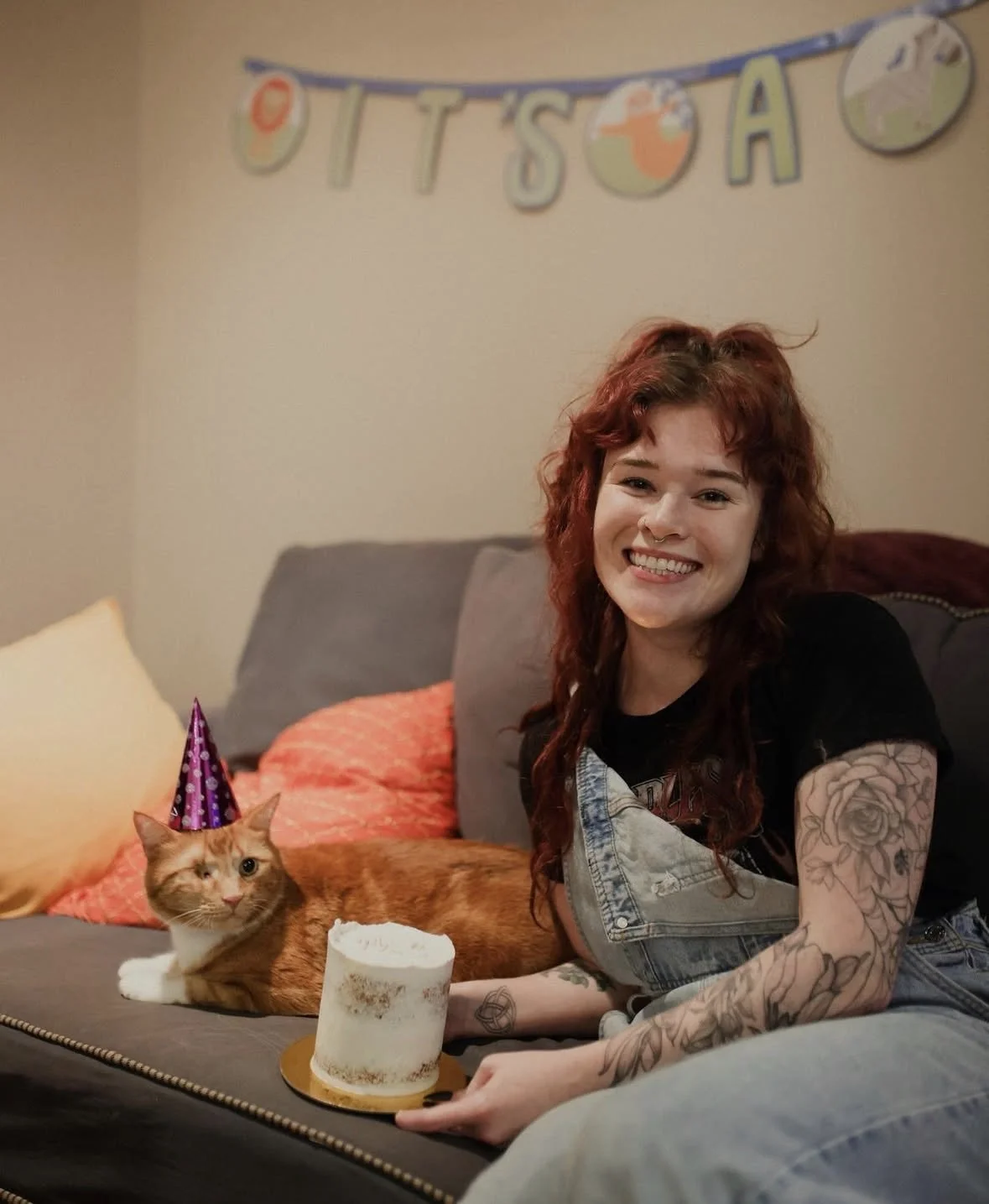 A young woman with red curly hair, tattoos on her arms, smiling and holding a small birthday cake, sitting on a gray couch with a ginger cat wearing a purple party hat next to her. In the background, a colorful banner spells out "OTTSO" and there are pillows on the couch.