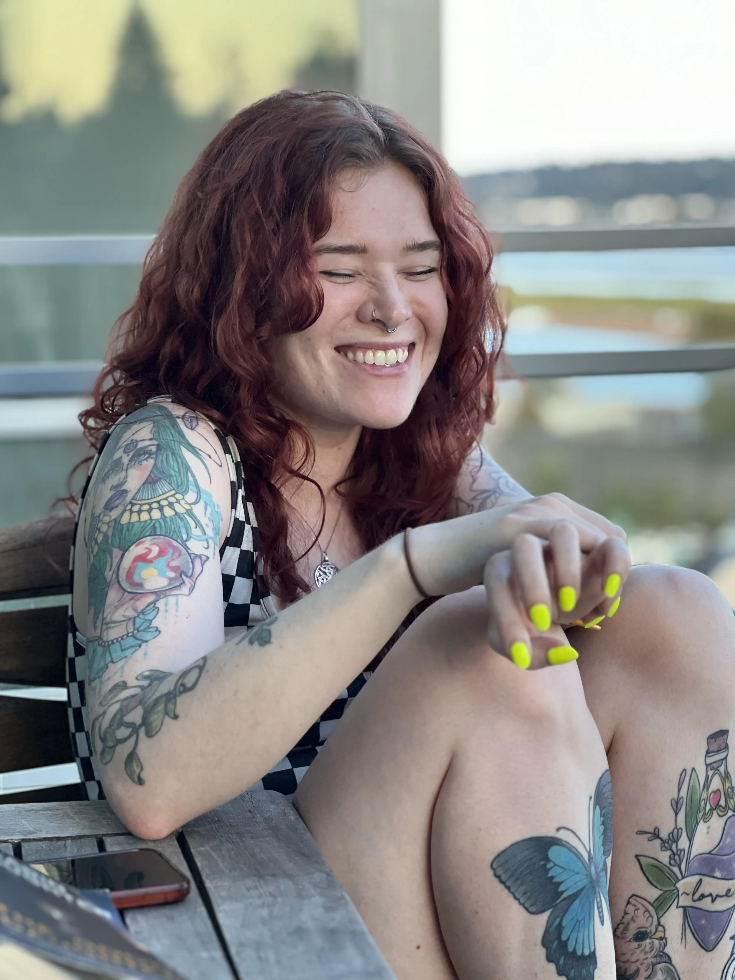A young woman with red curly hair and tattoos on her arms and legs, smiling and sitting outdoors on a wooden bench.