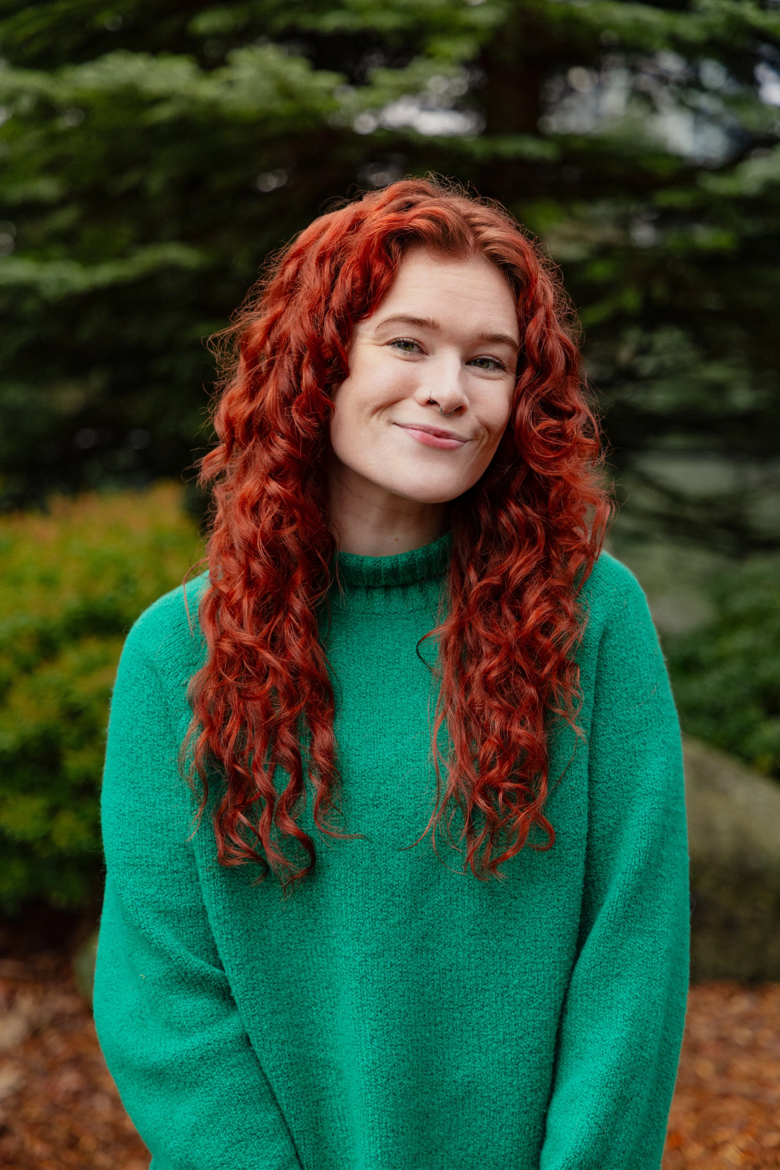 A woman with long, curly red hair and a nose piercing, wearing a green sweater, standing outdoors with a background of greenery.