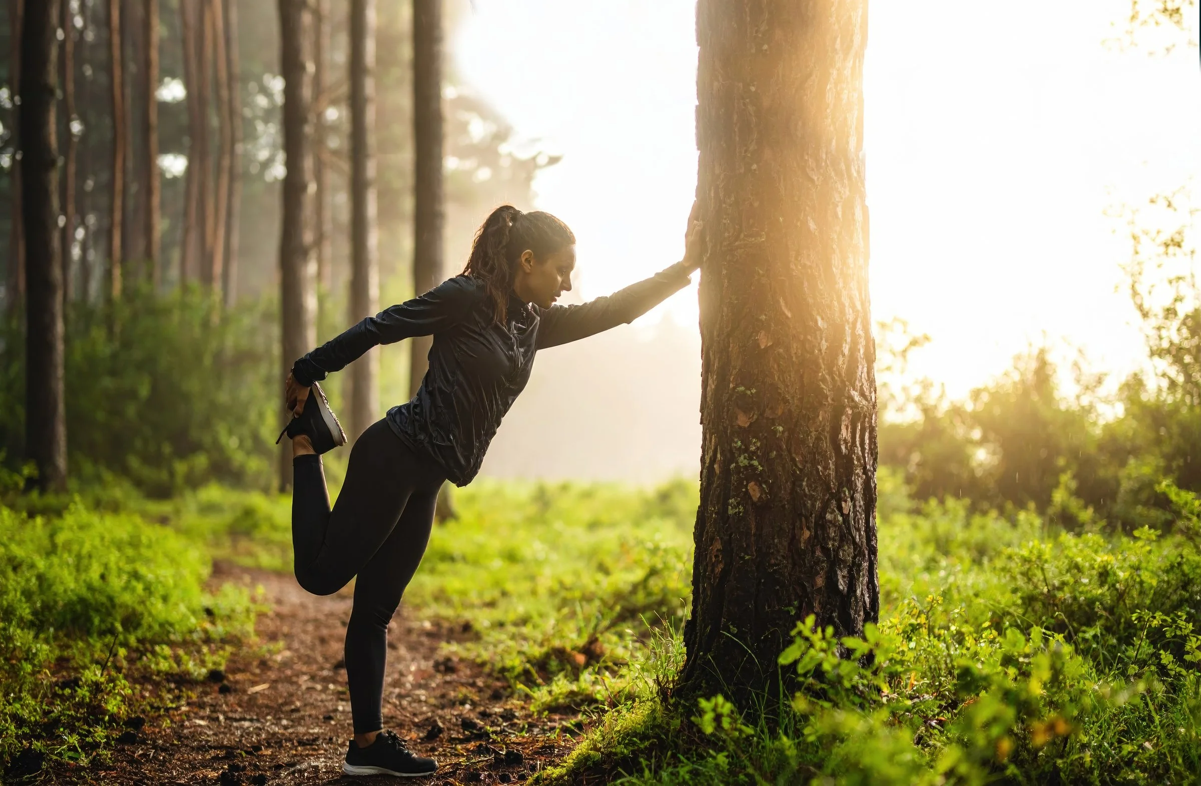 A woman doing a stretch exercise in a forest during sunrise or sunset, with one leg bent behind and held by her hand, and the other leg straight on the ground.