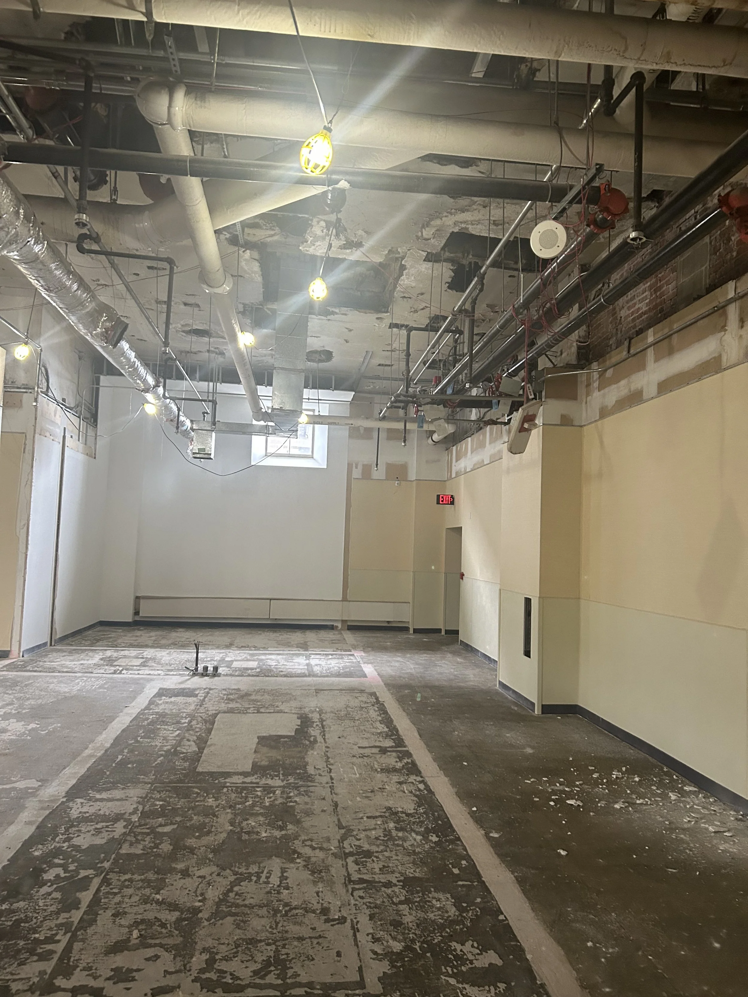 An unfinished interior space with exposed pipes on the ceiling, a partially removed ceiling surface, and a damaged floor with debris, paint marks, and markings for construction or renovation.