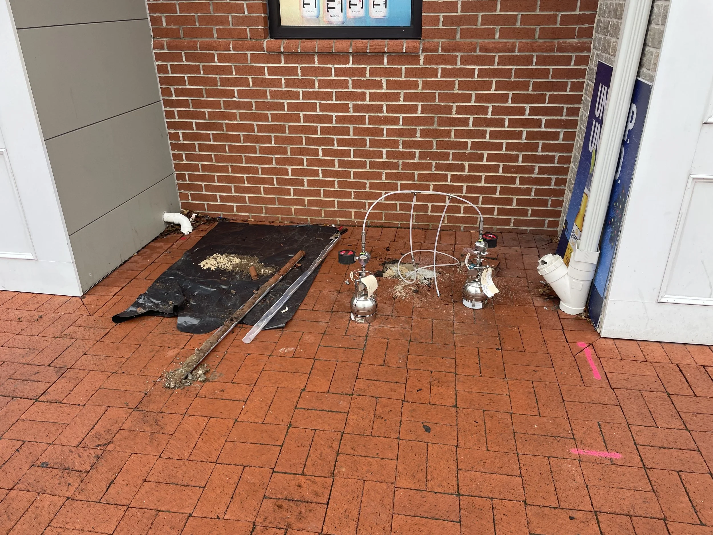 A brick wall with sub-slab vapor equipment on a brick patio. There are two sample canisters, sample tubing, a black tarp with debris, and some tools on the ground.