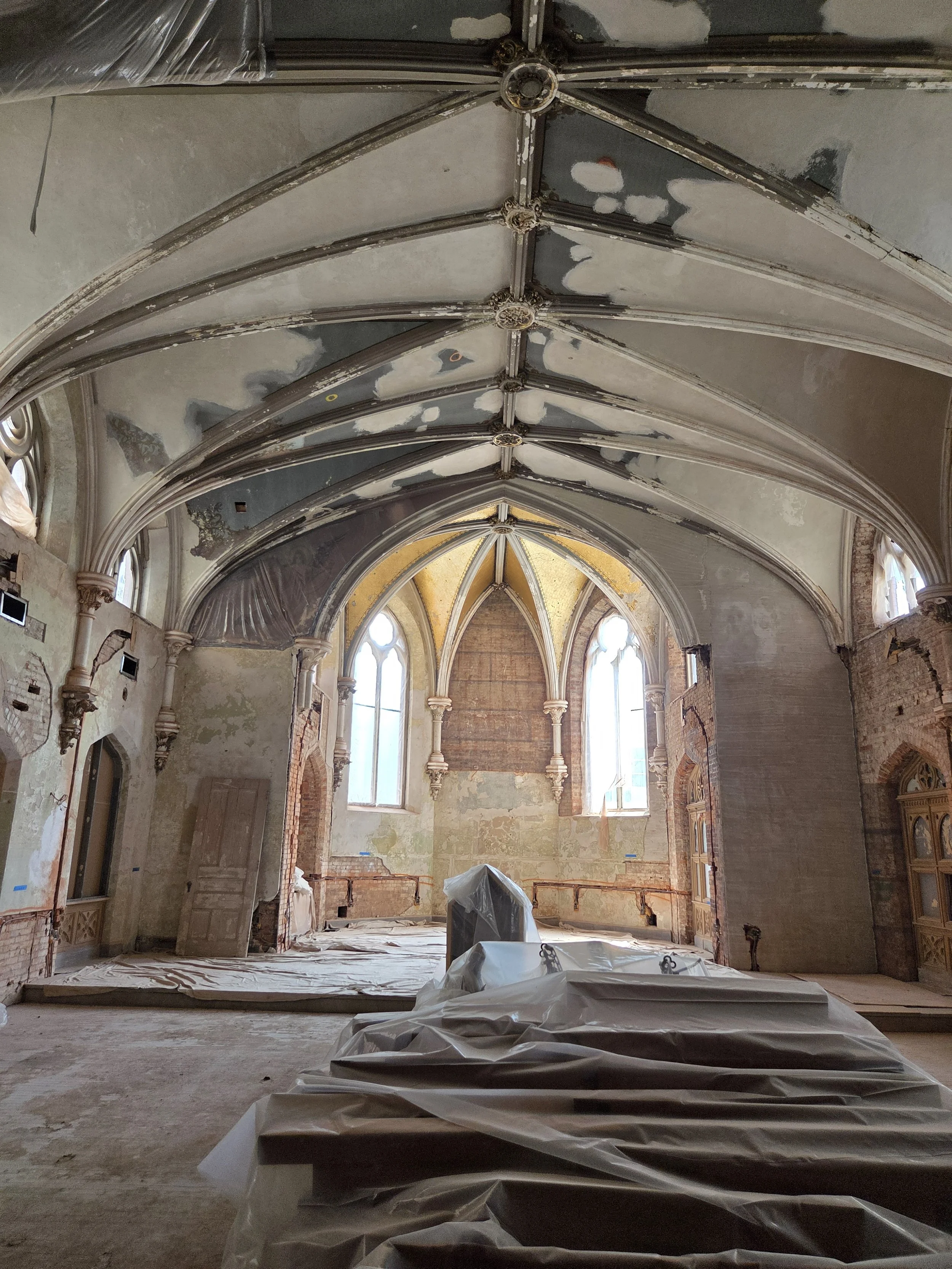 The interior of a church or cathedral undergoing renovation, with high vaulted ceilings, arched windows, exposed brick walls, and construction materials covered in plastic sheeting on the floor.