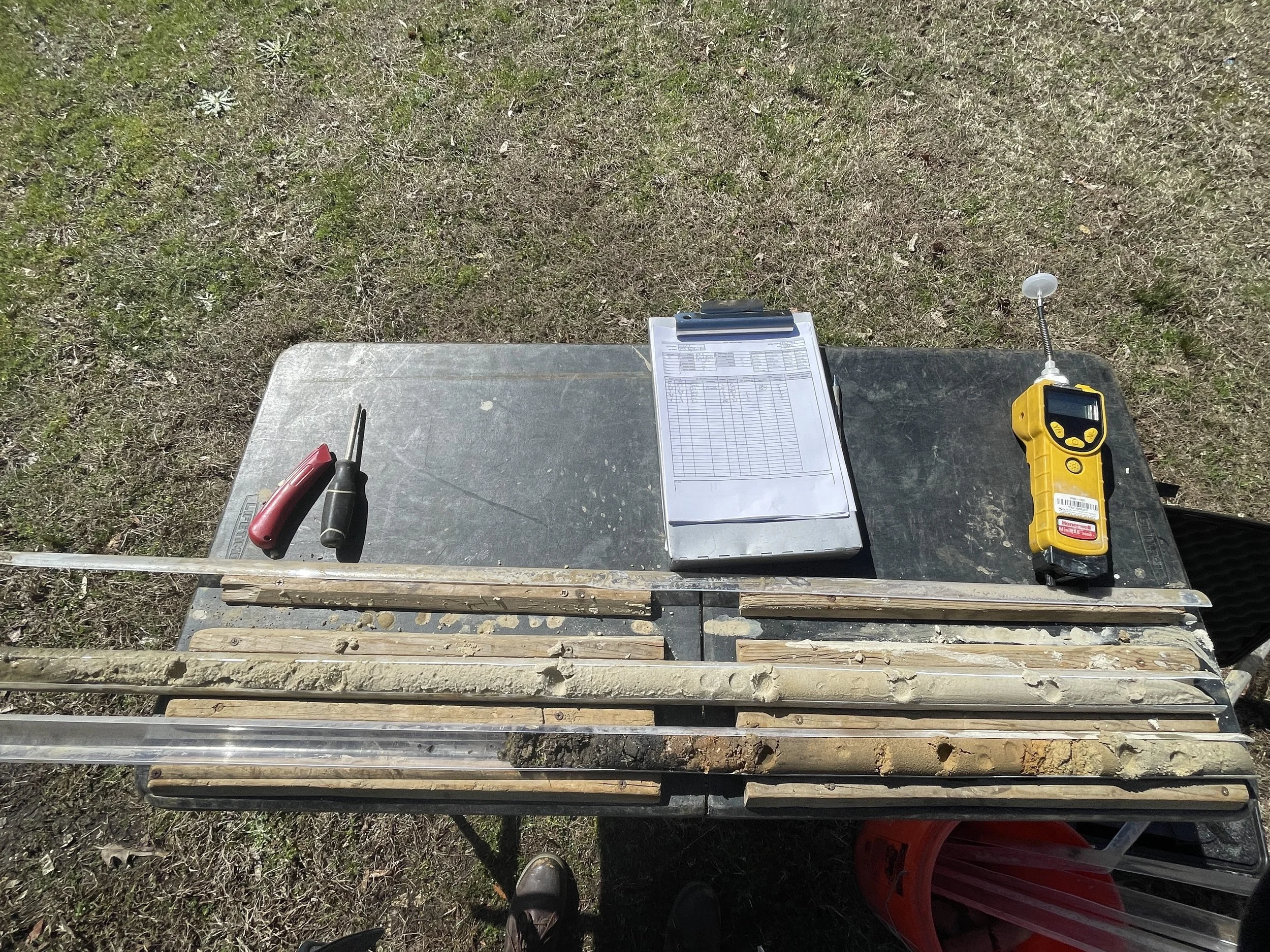 A flat surface with tools and equipment, including a clipboard with papers, a small yellow electronic device, screwdrivers, and a long level, set on a metal and wooden framework with grass in the background.