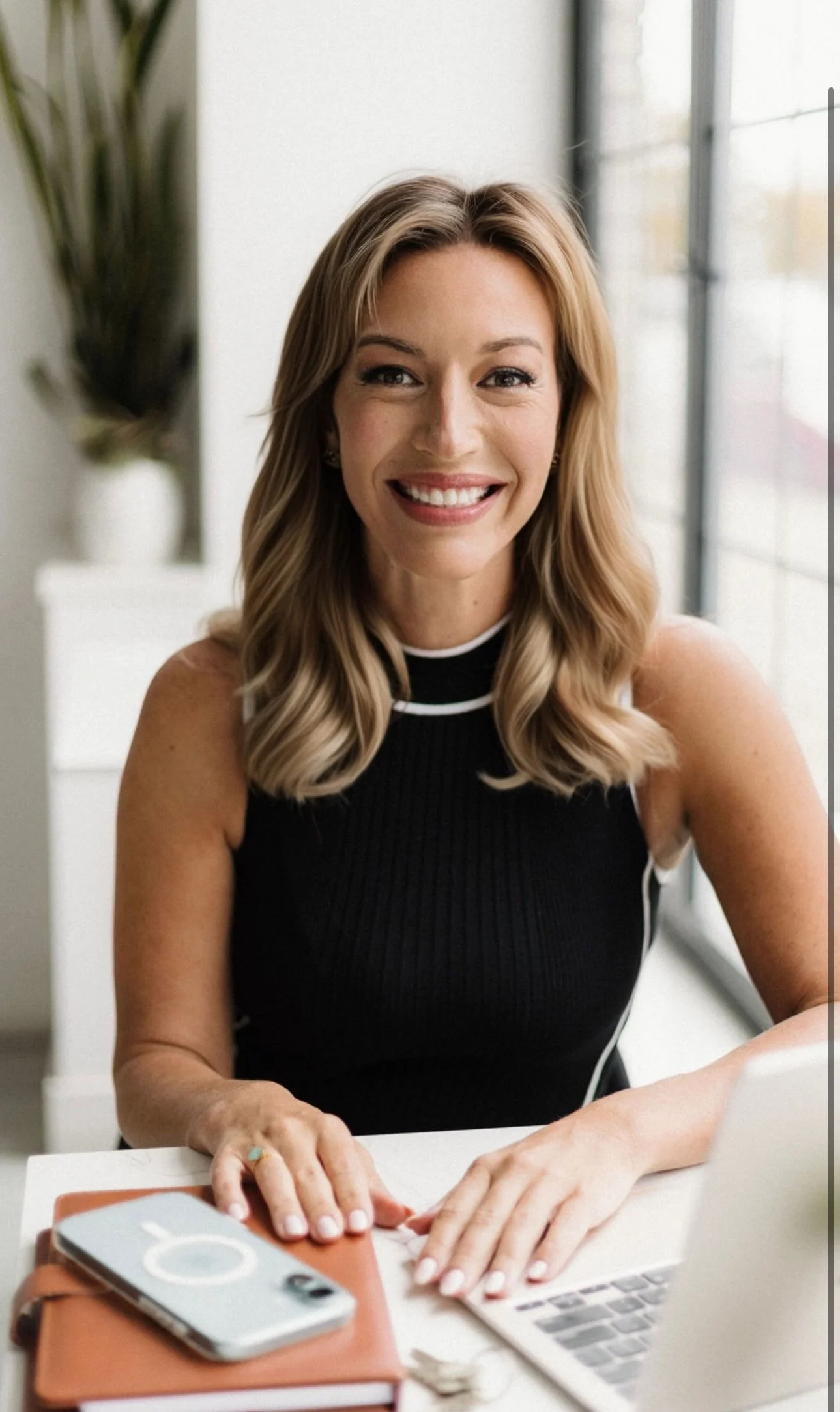 A woman with blonde hair smiling at the camera, sitting at a desk with a laptop, phone, and notebooks in a bright room with large windows and a plant in the background.